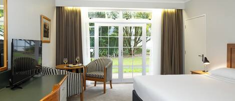 A hotel room with a bed, a desk with a chair, a television, and a view of the outdoors through large glass doors.