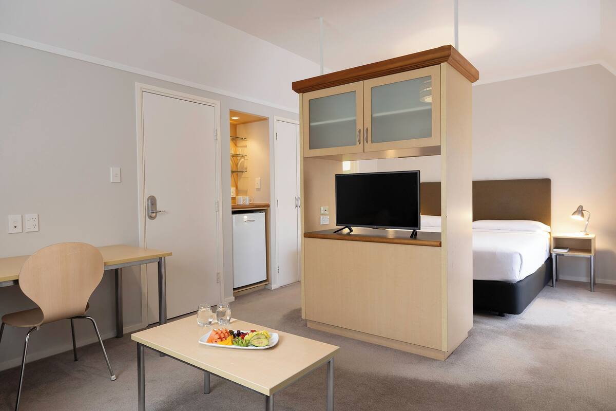 Een kleine hotelkamer met een bed, een televisie, een klein kitchenette, een bureau en een stoel.