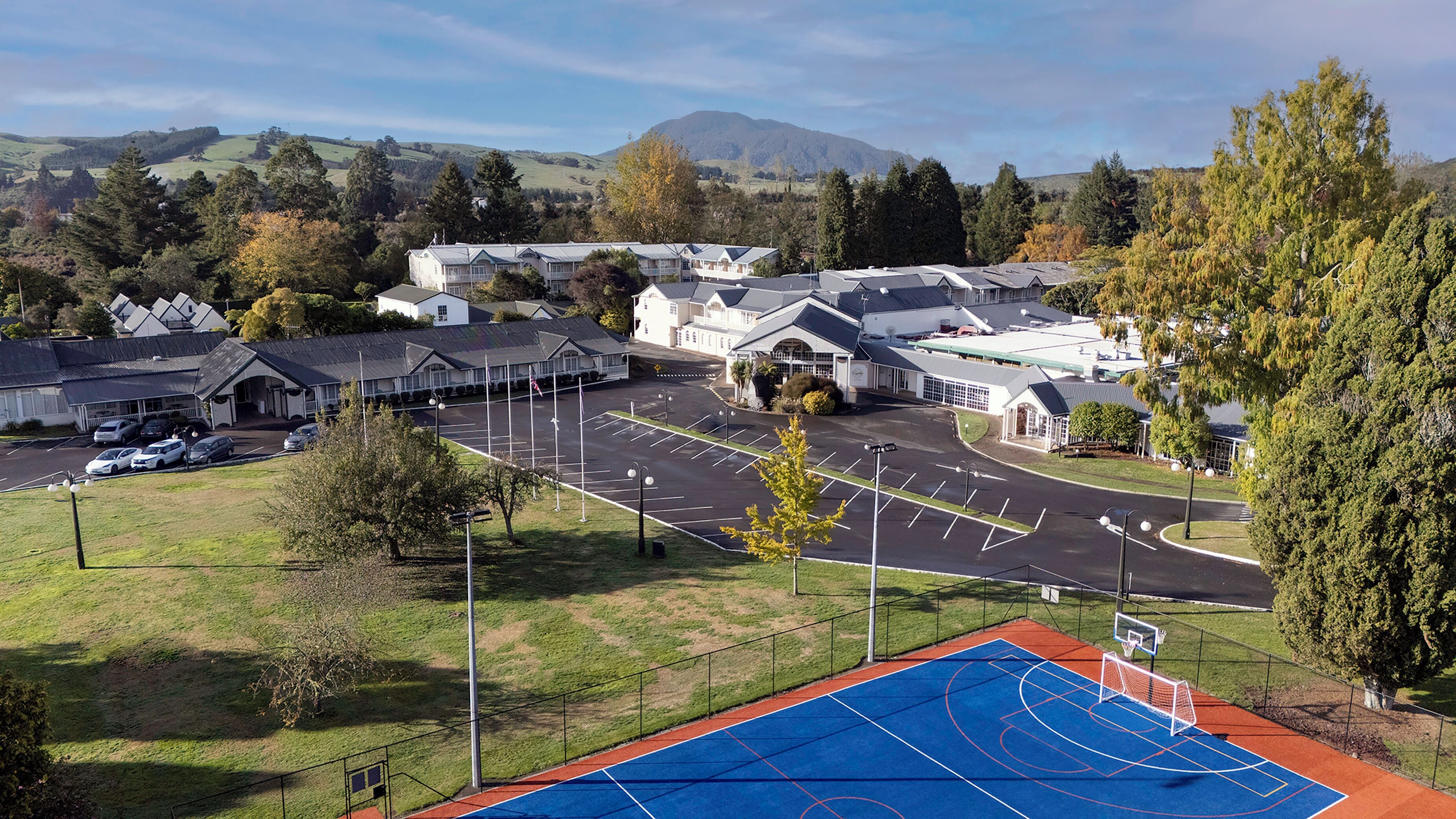 A sports court with a basketball hoop, a parking lot, and buildings in the background.