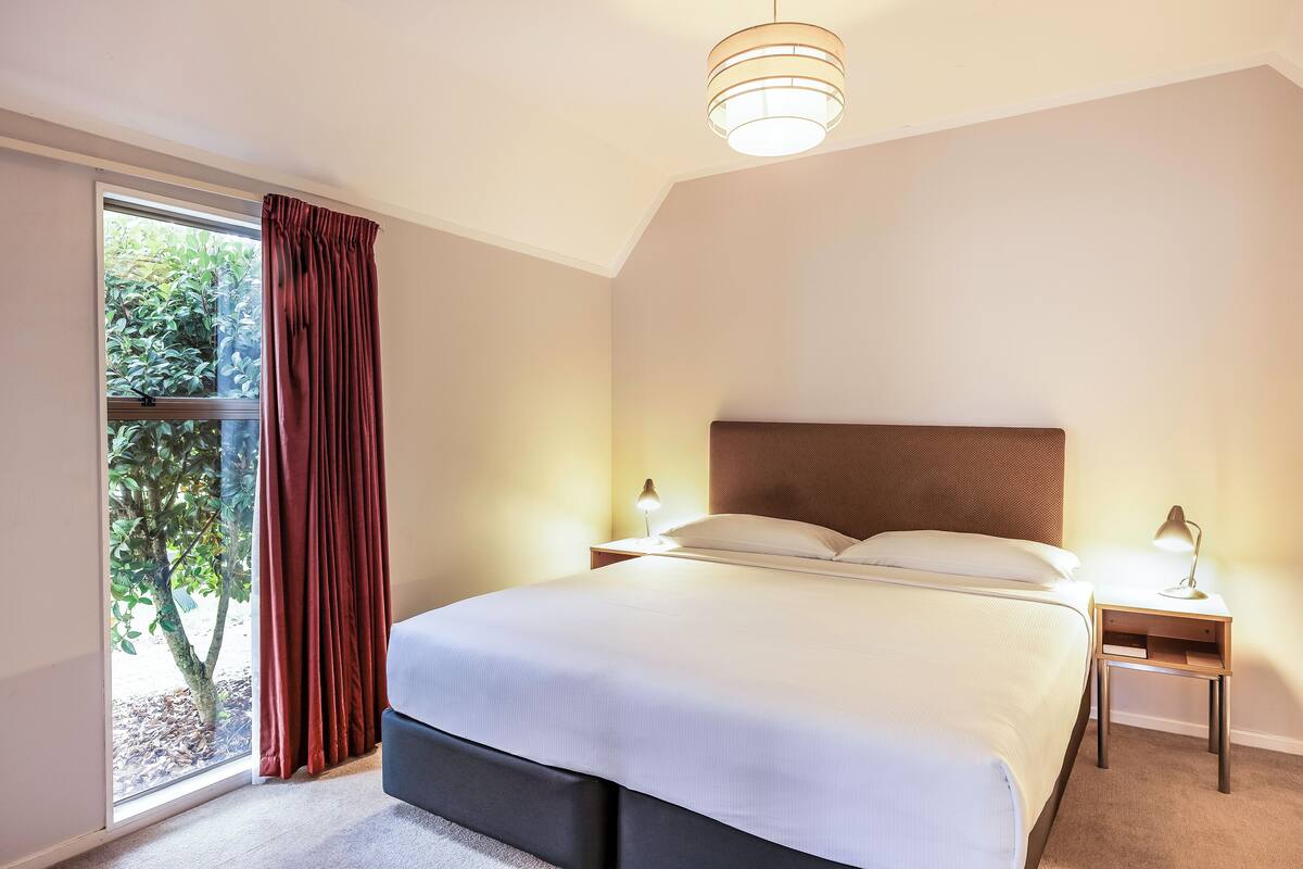 Een netjes opgemaakt bed in een kamer met een groot raam, een nachtkastje met een lamp en een plafondarmatuur.