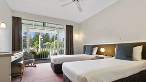 Habitación de hotel con dos camas, un escritorio y un balcón que da a árboles y una bandera.