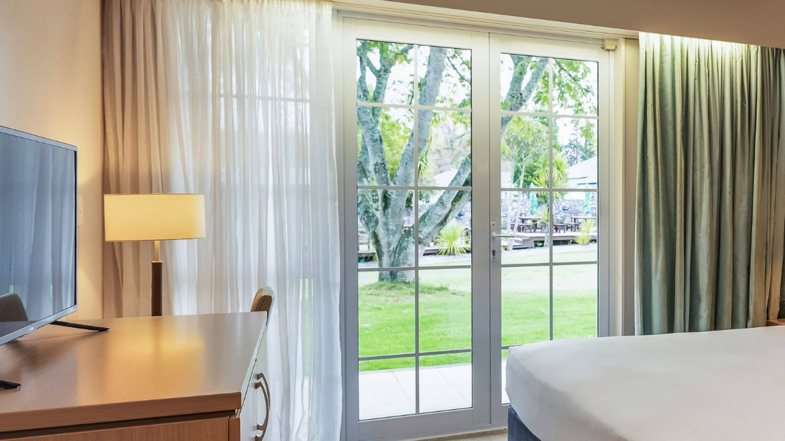 A hotel room with a bed, a desk with a lamp, a television, and a sliding glass door leading to a patio with a view of trees and a building.