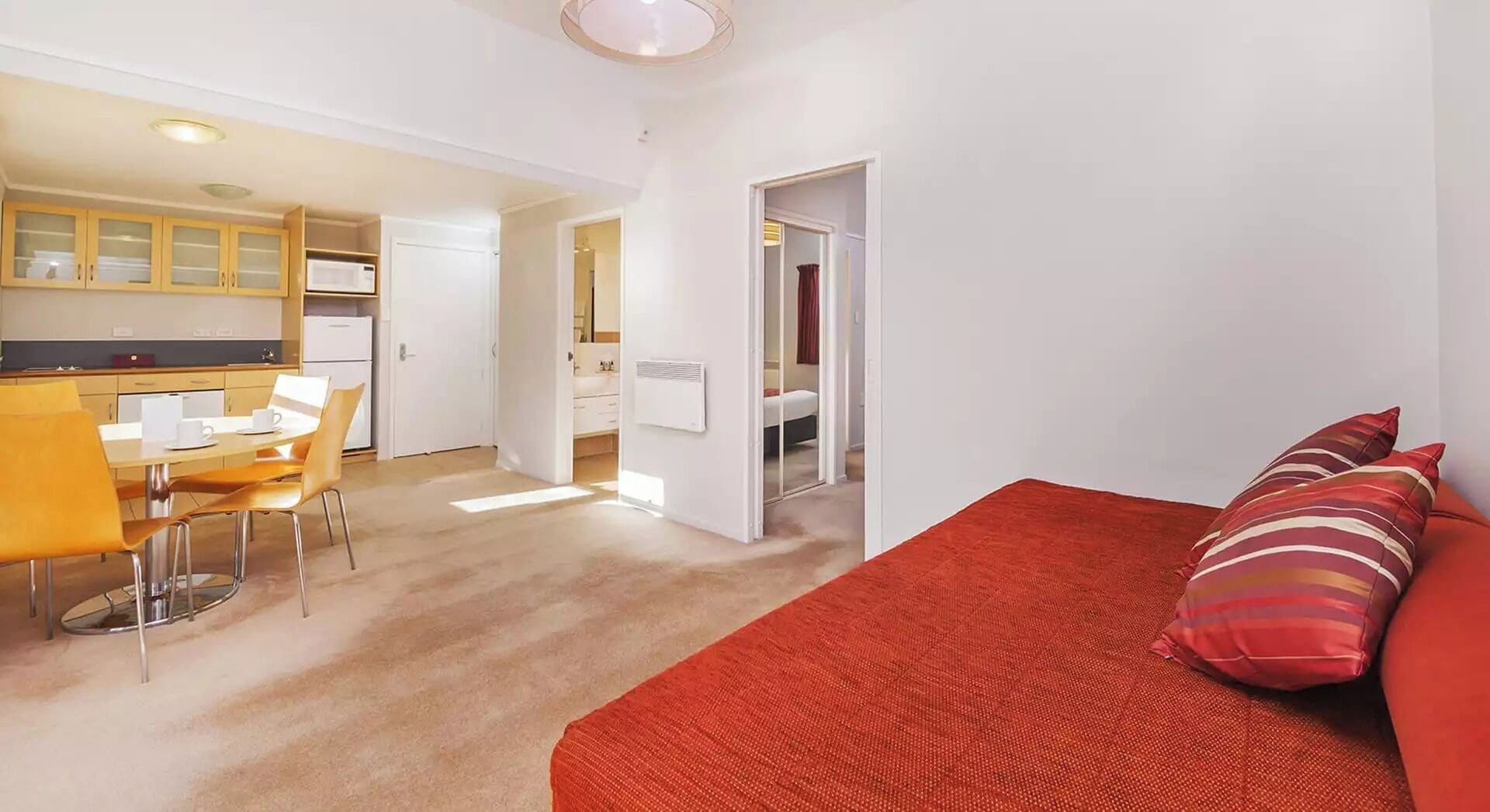 Una habitación de hotel compacta con un sofá rojo, un área de comedor con mesa y sillas, y una kitchenette.