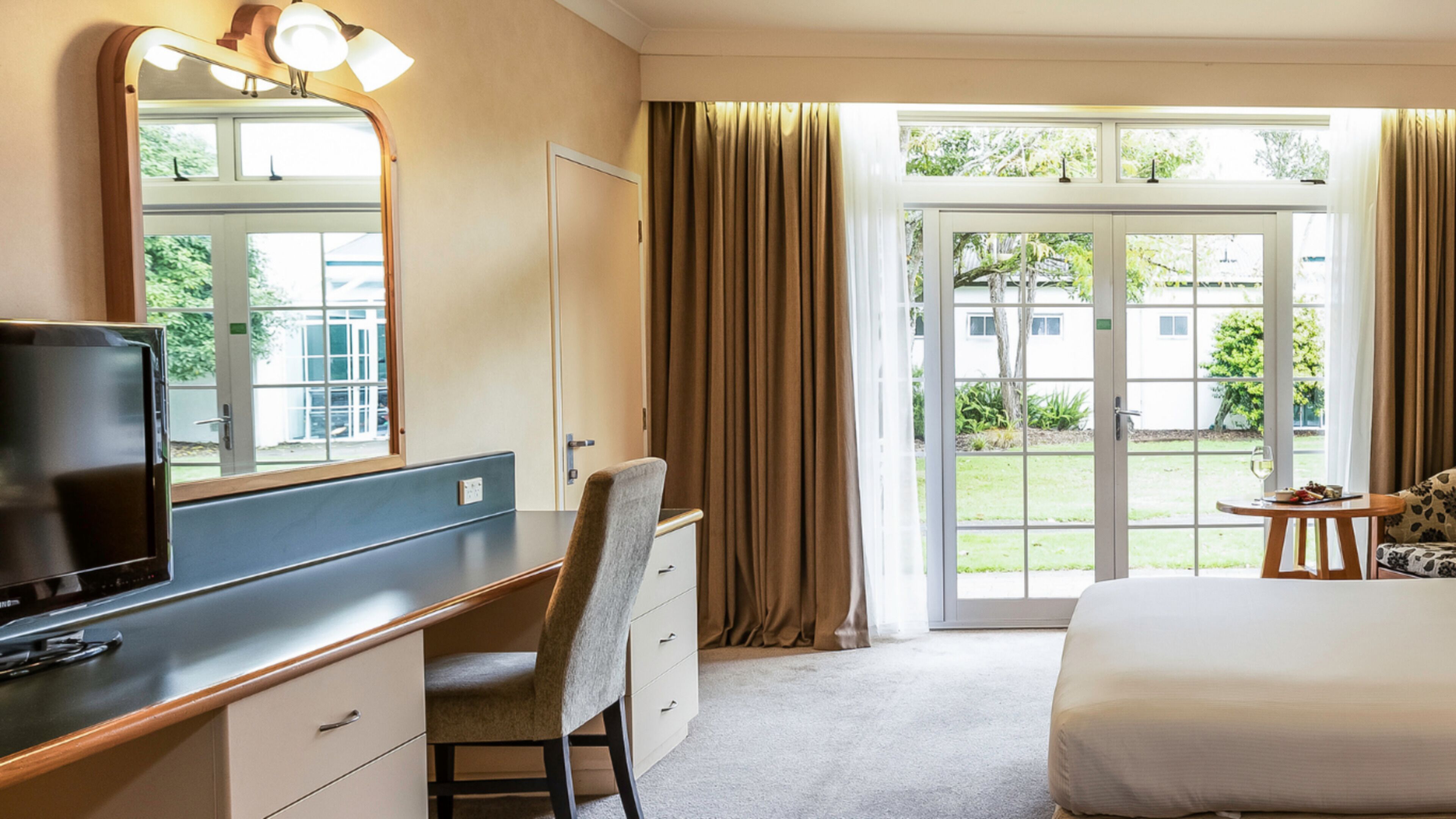 A hotel room with a bed, a desk with a chair, a television, a large mirror, and a view of the outdoors through a glass door.