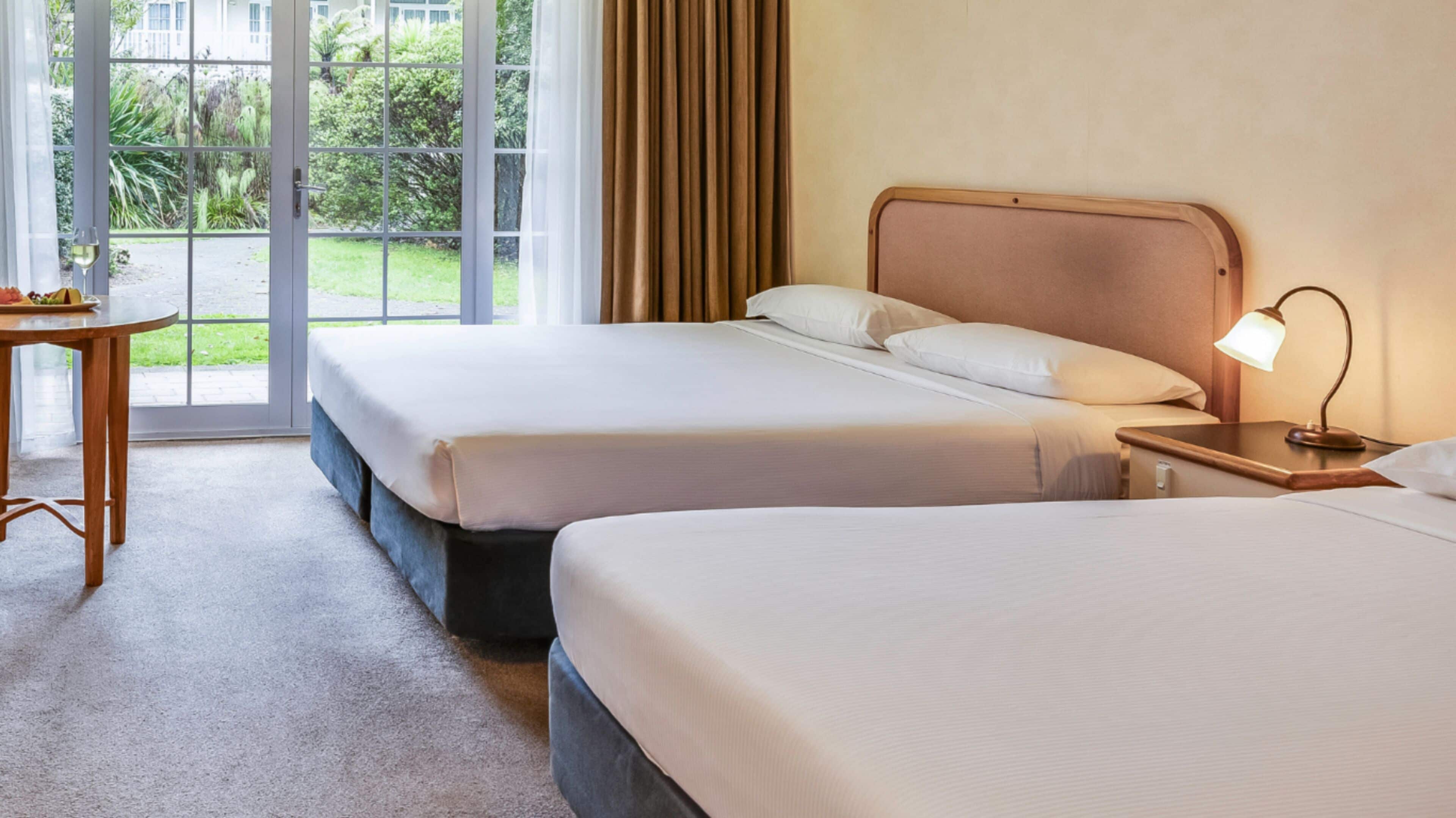 A hotel room with two beds, a desk, and a view of greenery outside.