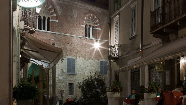 A narrow street with buildings on either side, a sign for 'Albergo Orlogio', and outdoor seating with potted plants. - Albergo Orologio (Brescia)