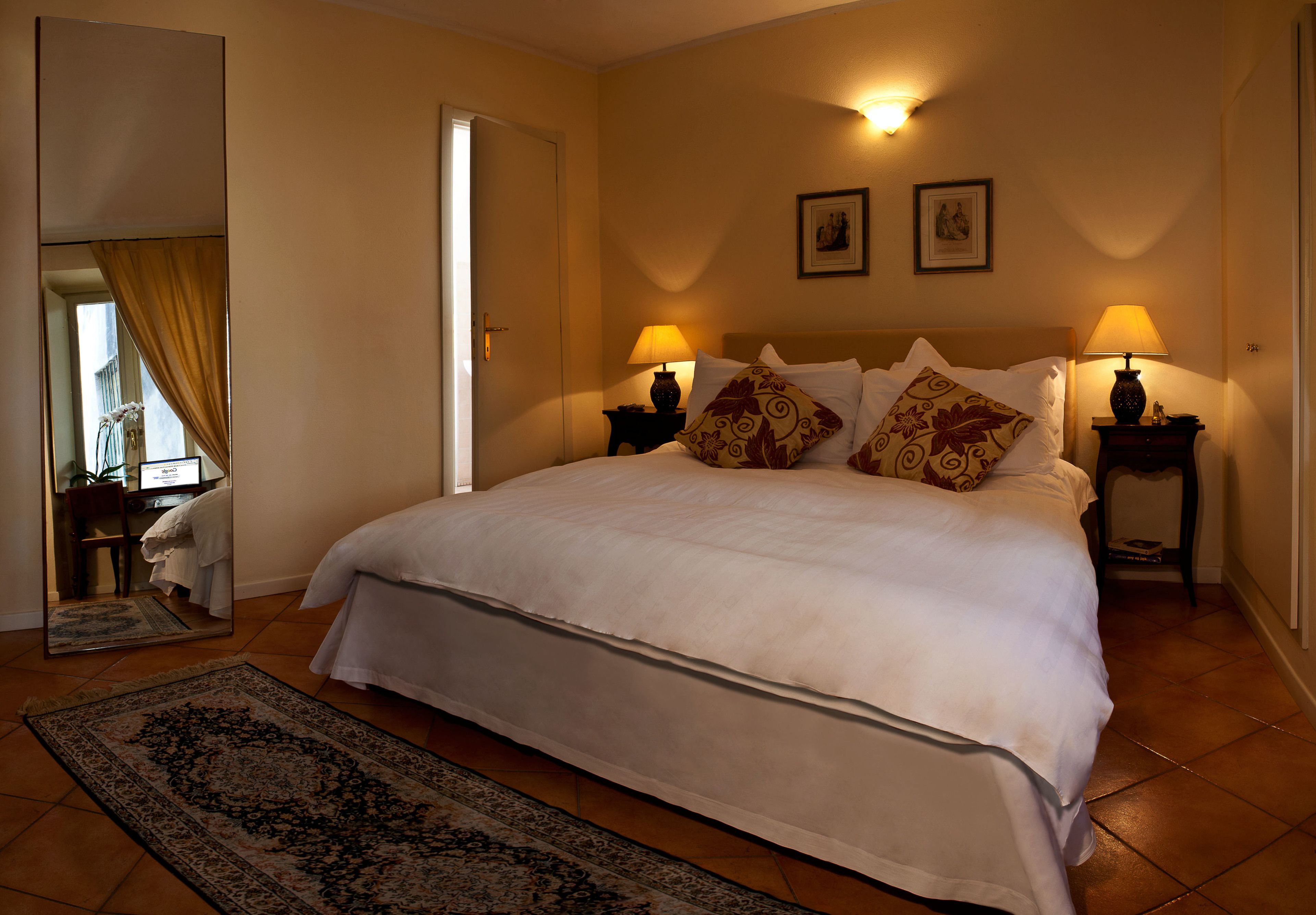 Una cama bien tendida con sábanas blancas y almohadones decorativos, dos lámparas de noche y un espejo grande que refleja parte de la habitación.