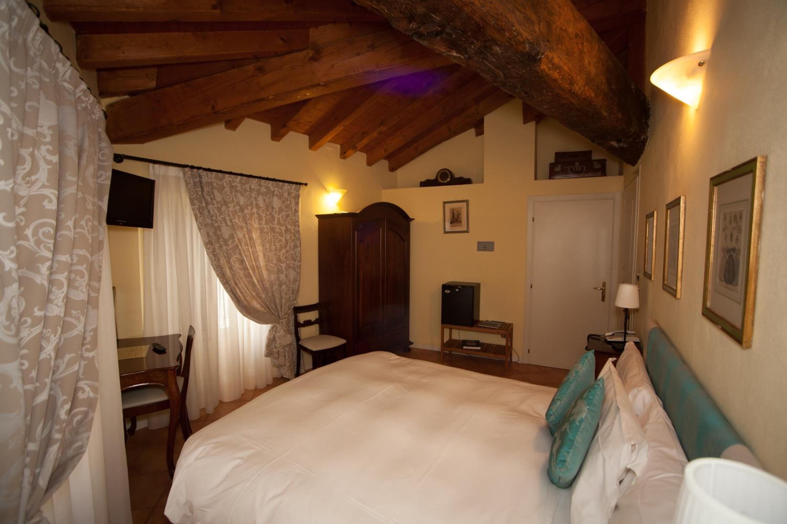 A bedroom with a large bed, wooden ceiling beams, a desk with a chair, a wardrobe, and a television.