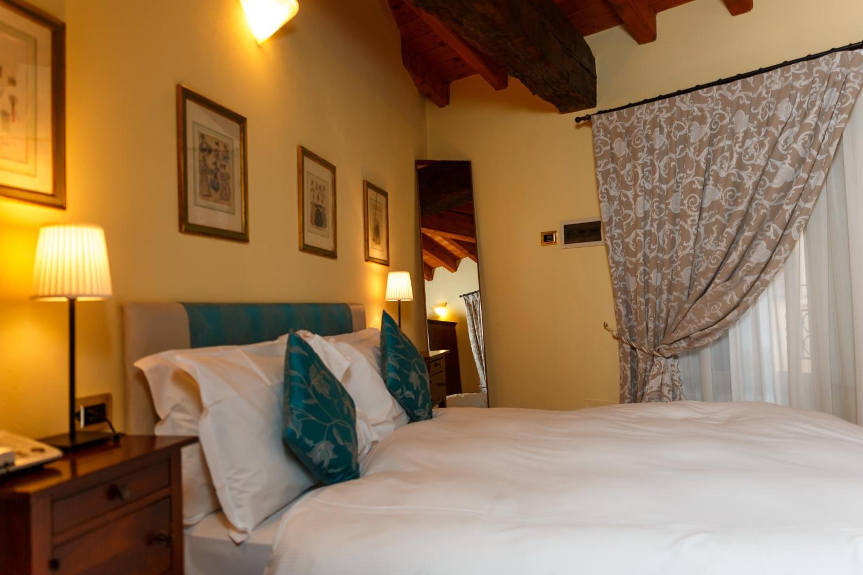 Una camera d'albergo con un letto, un comodino, una lampada, quadri appesi al muro e una finestra con tende.