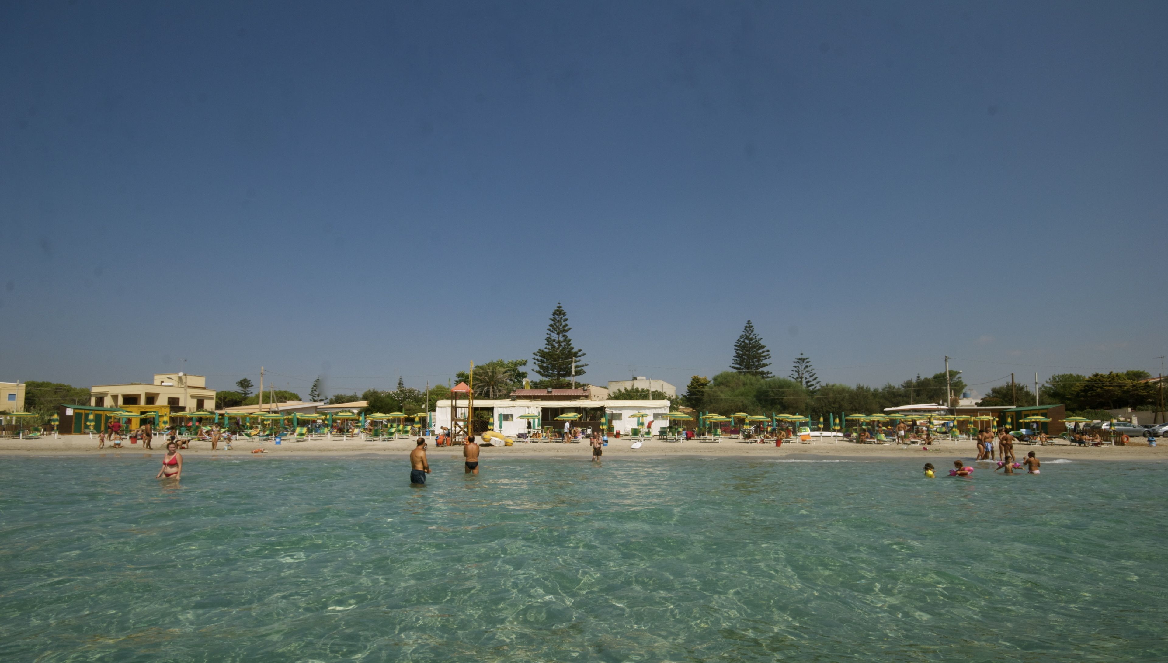 Een strand met helder water, ligbedden, parasols en enkele mensen.