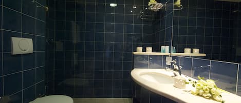 A bathroom with dark blue tiles, a white toilet, a sink with a mirror, and a shelf with toiletries.