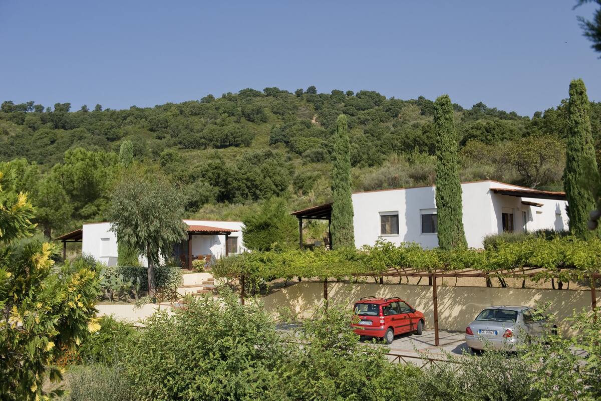 A hillside property with white buildings, a red car, and a white car parked in front.