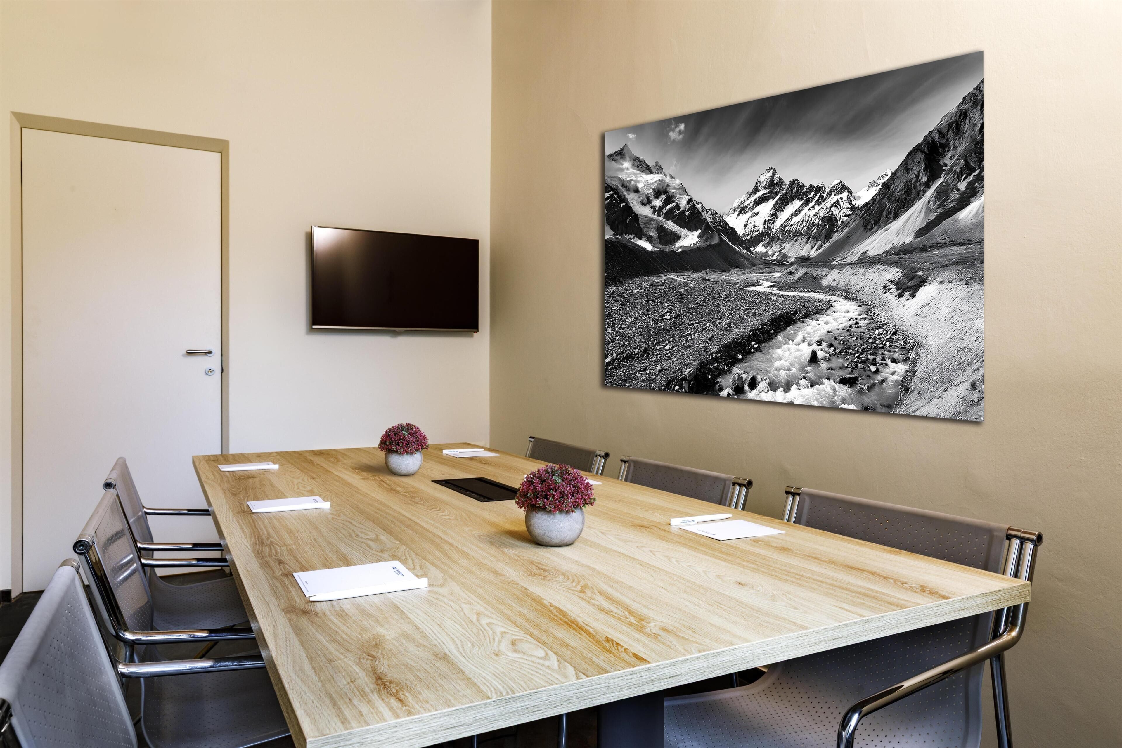 A conference room with a long wooden table, six chairs, two potted plants, a flat-screen TV, and a mountain landscape painting on the wall.