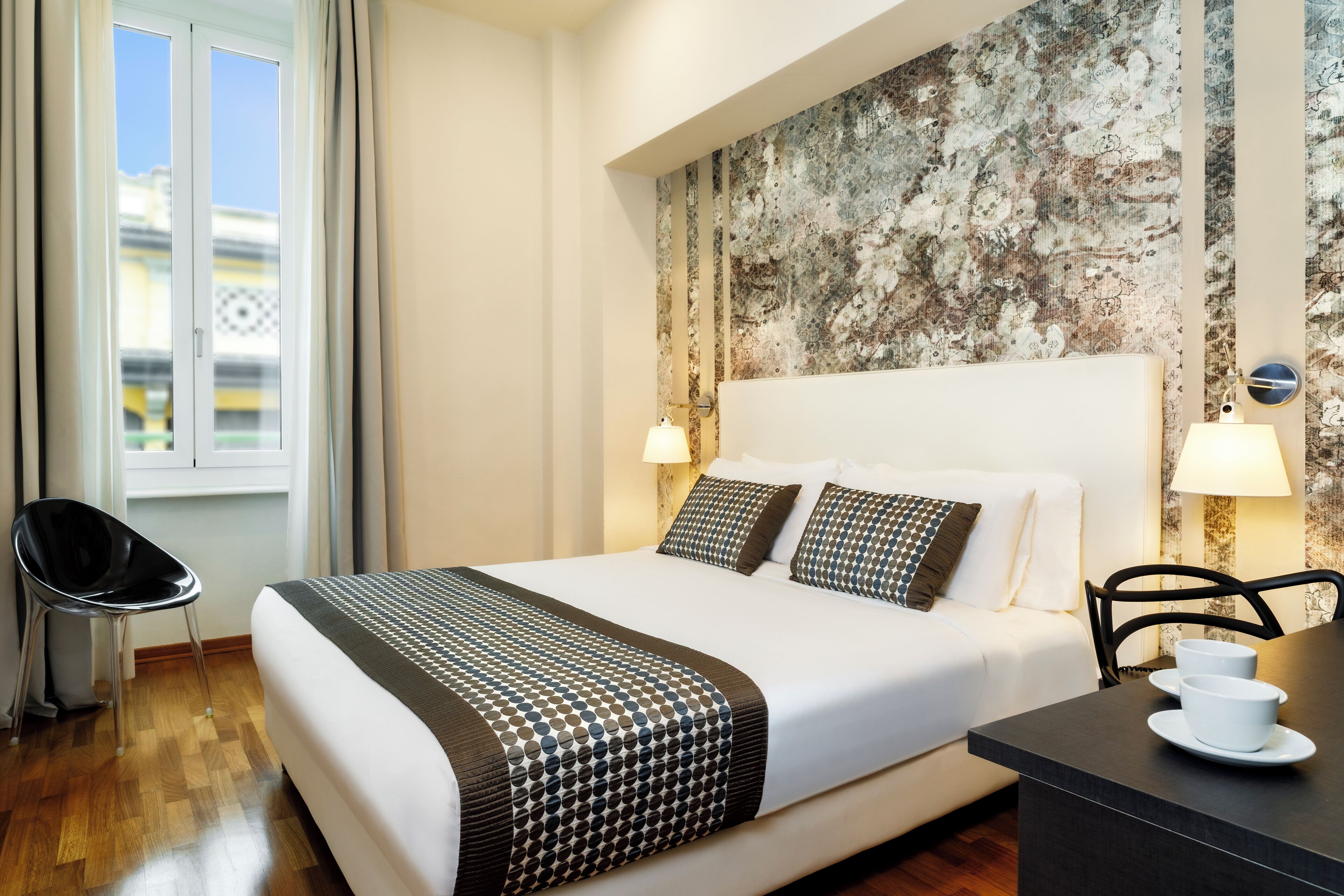 A hotel room with a large bed, two bedside lamps, a small table with cups, and a chair.
