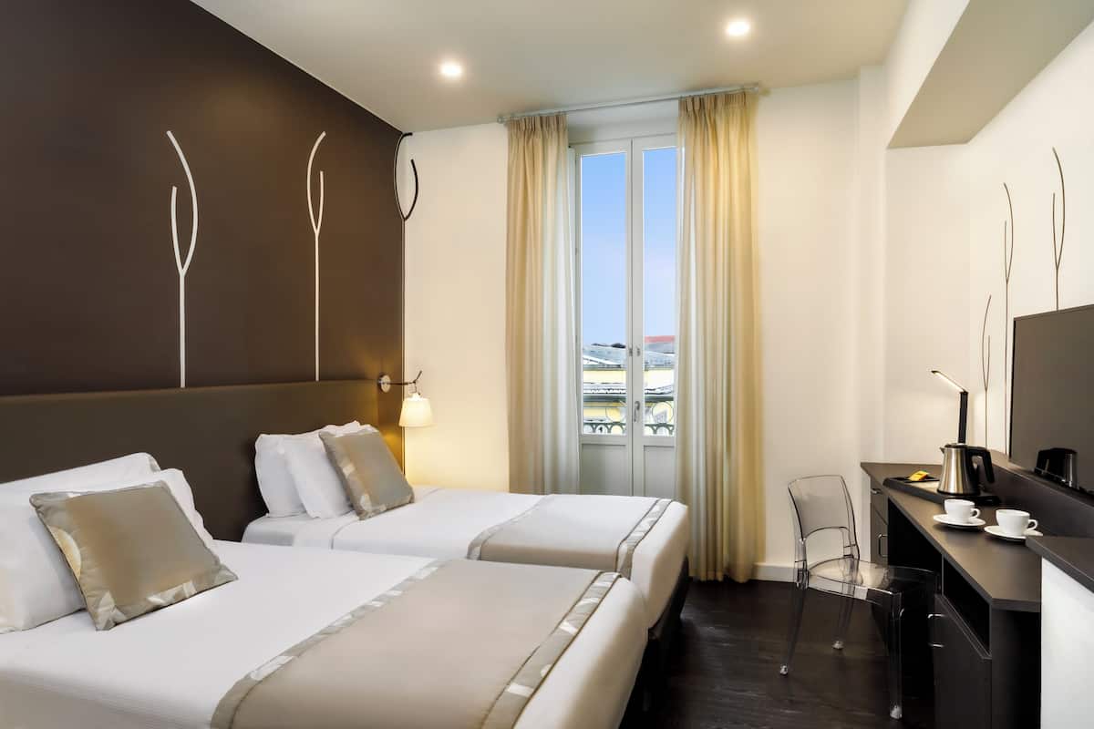 A hotel room with two beds, a desk, a chair, and a large window with curtains.