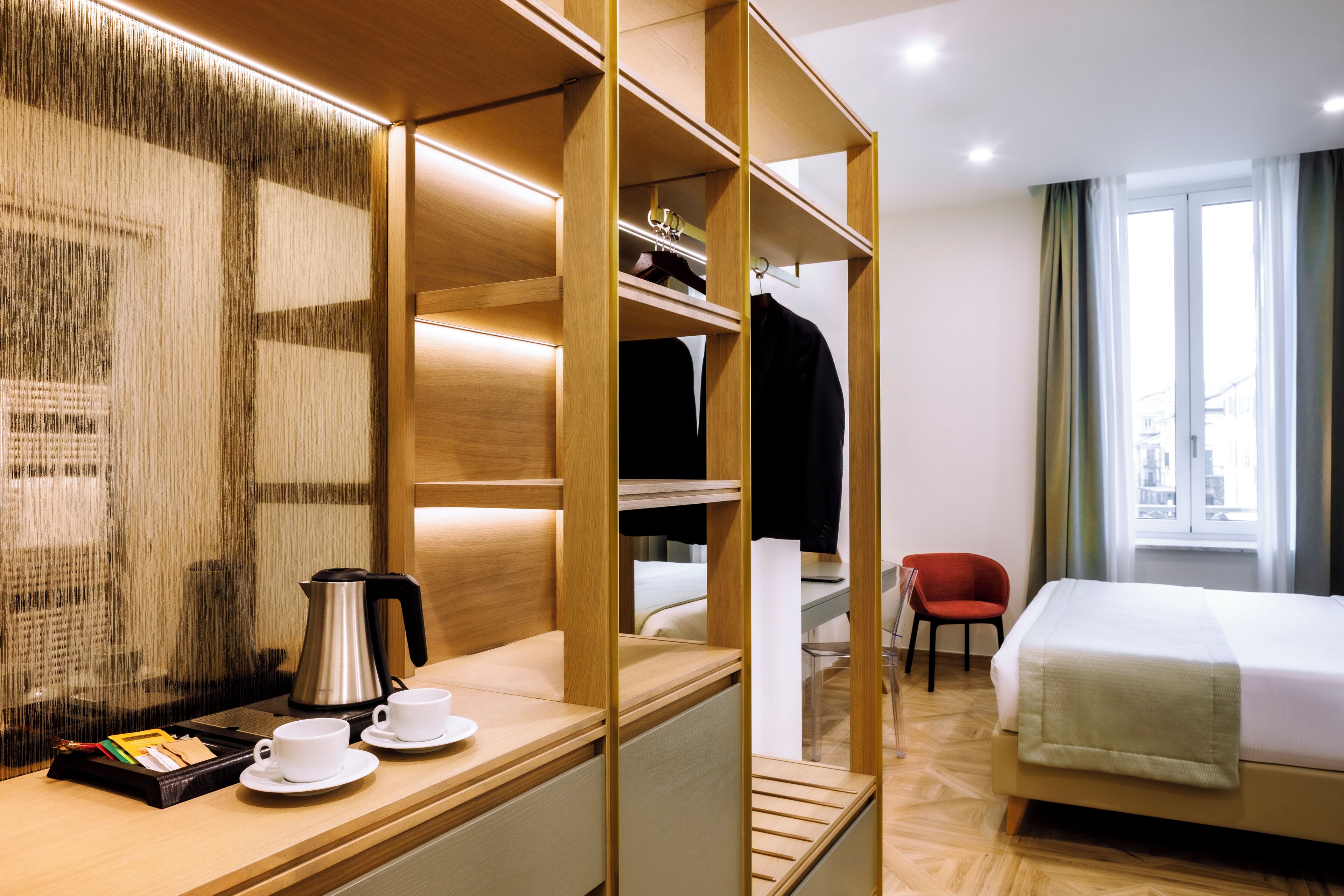 A hotel room with a wooden wardrobe, a bed, a desk with a coffee maker, cups, and a chair.