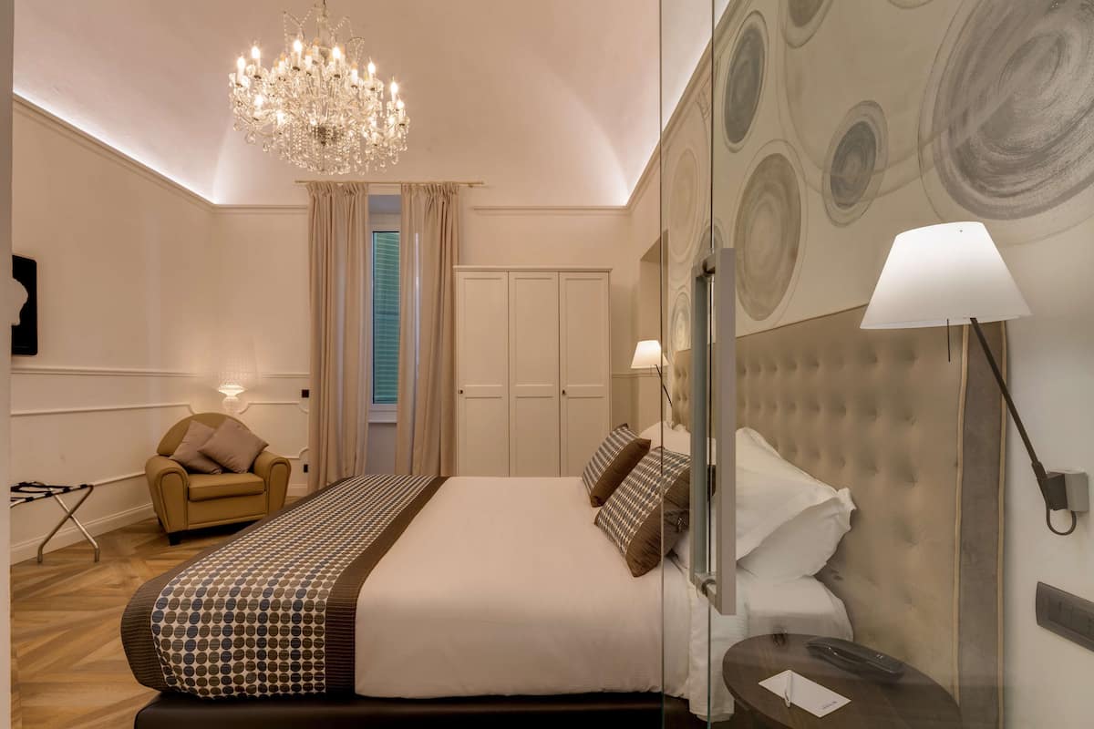 A hotel room with a large bed, a chandelier, a sofa, and a mirror with a lamp.