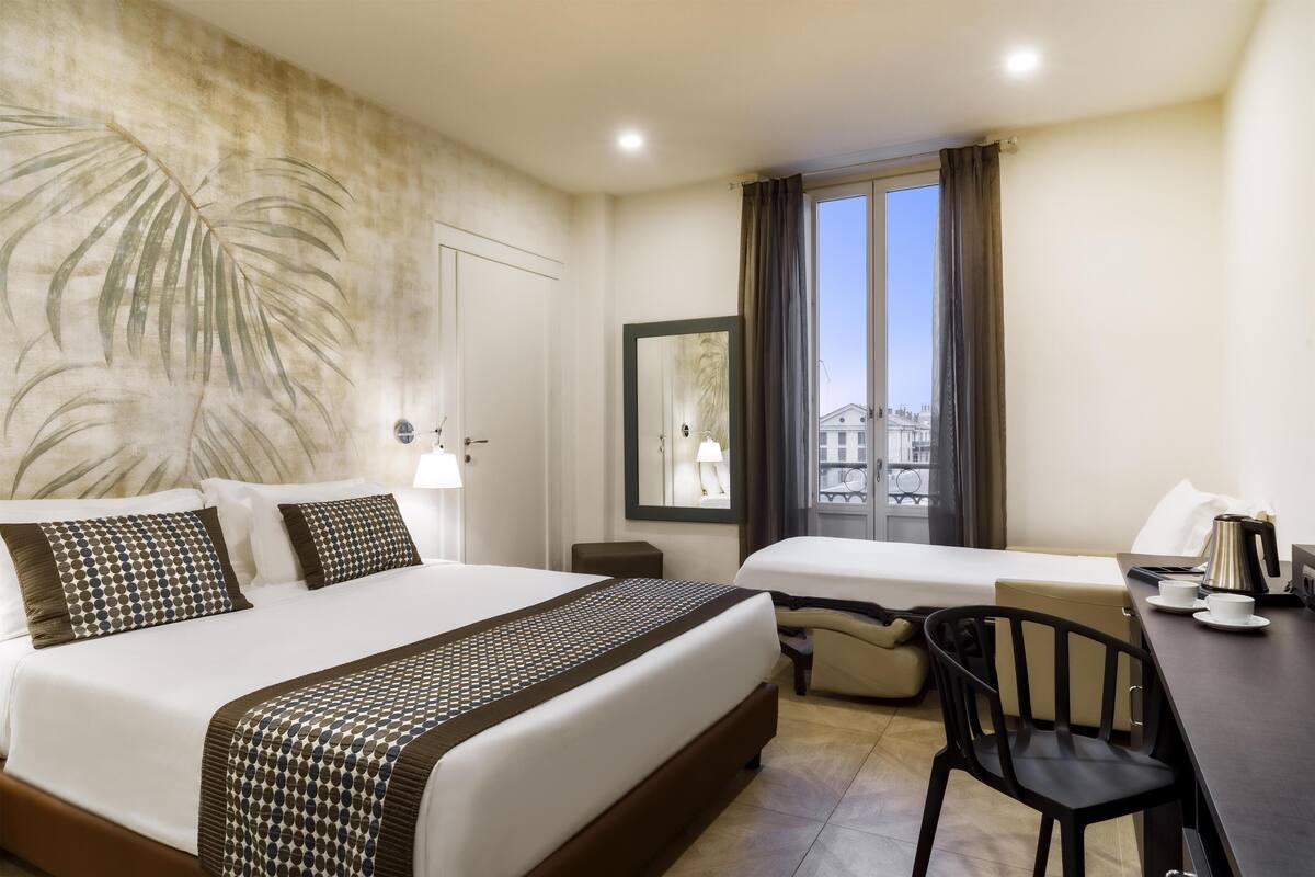 A hotel room with a large bed, a smaller bed, a desk with a chair, and a view of buildings through a window.
