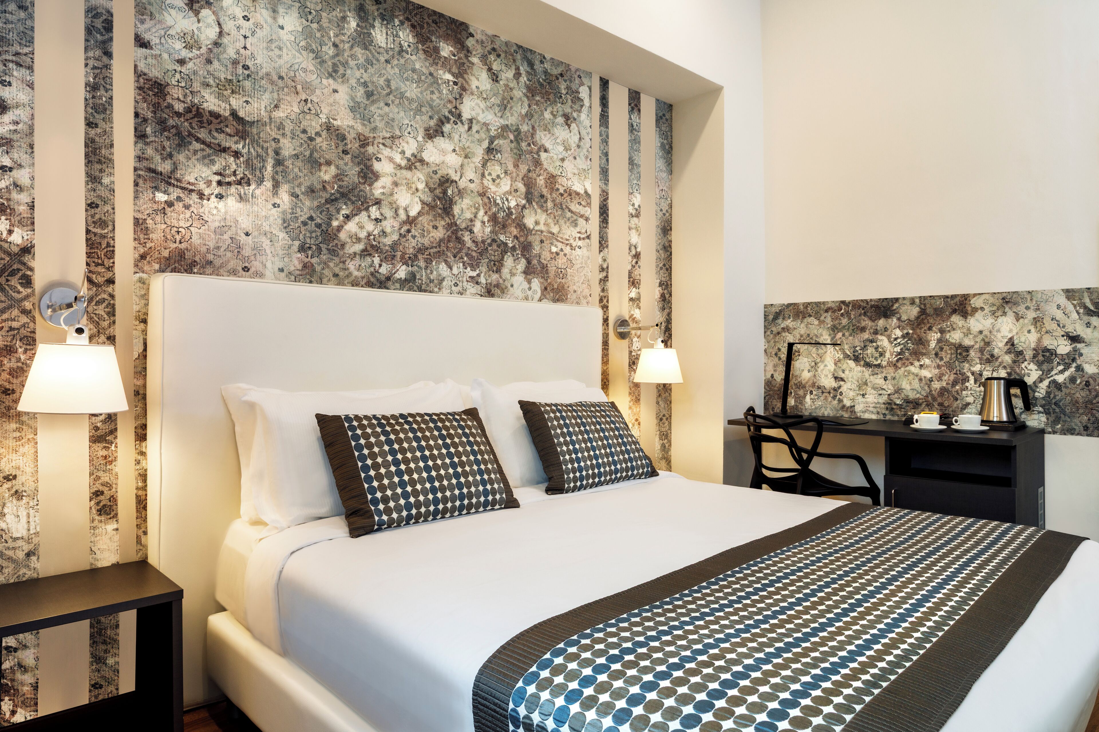 Una habitación de hotel con una cama grande, dos lámparas de noche, un escritorio con cafetera y una pared con diseño.