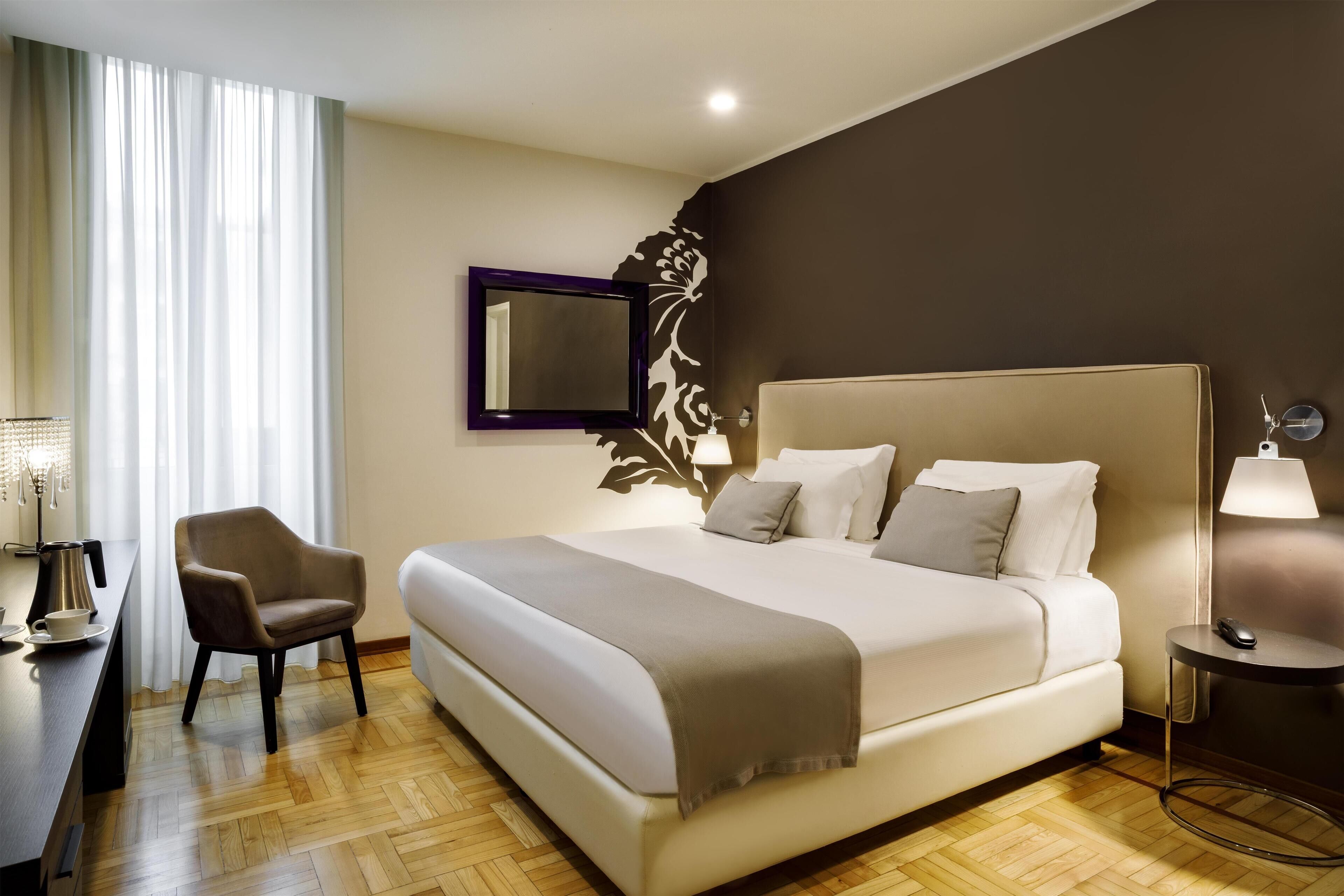 A hotel room with a large bed, a chair, a desk with a lamp, and a wall-mounted mirror.