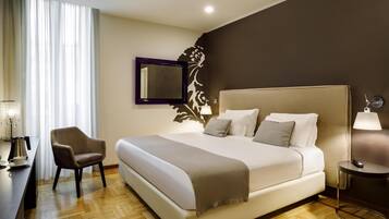 A hotel room with a large bed, a chair, a desk with a lamp, and a wall-mounted mirror.