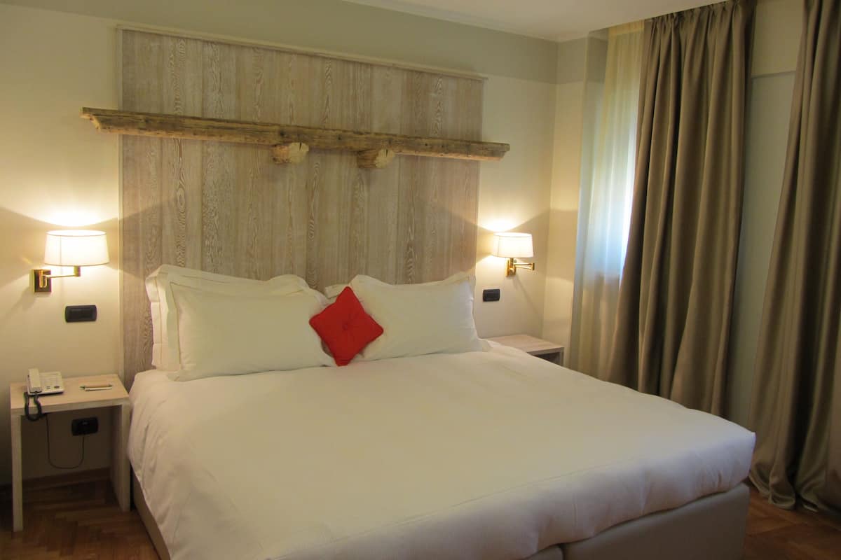 Habitación de hotel con una cama grande, dos mesitas de noche, una lámpara de noche, cabecero de madera, dos lámparas fijadas a la pared y una cortina.
