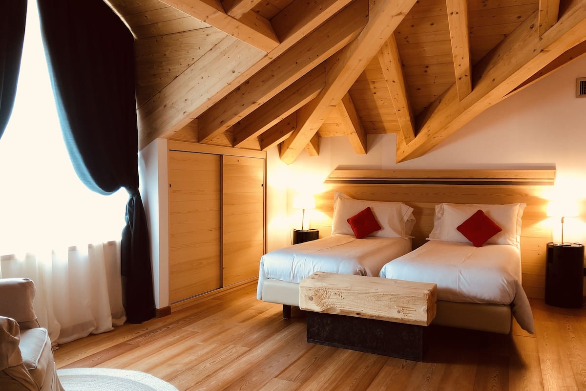 Un dormitorio con techo de madera, dos camas con ropa blanca y almohadas rojas, una mesita de noche con lámpara y una ventana con cortinas.
