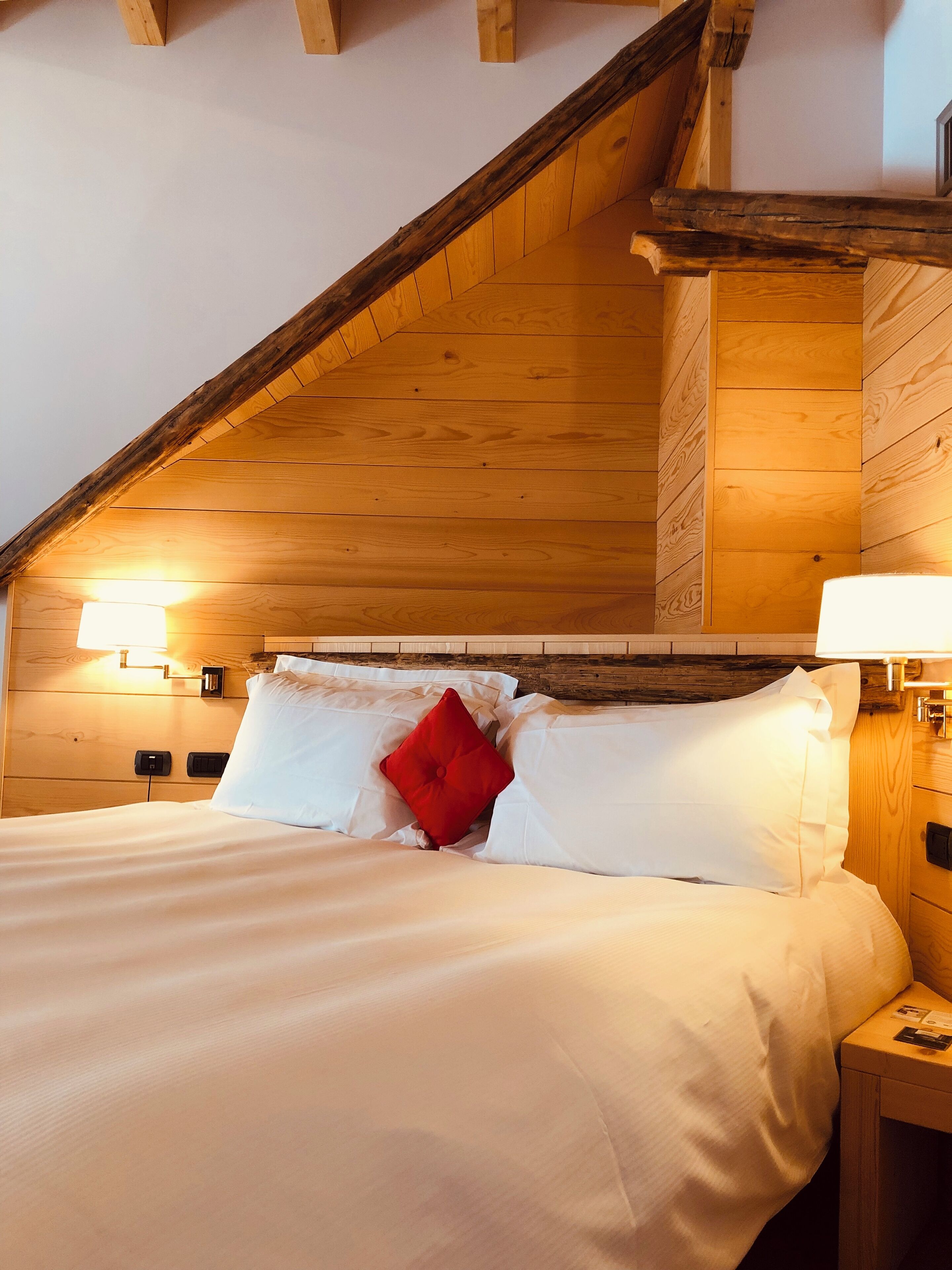 A bedroom with a sloped ceiling, wooden walls, a bed with white bedding and a red pillow, and bedside lamps.