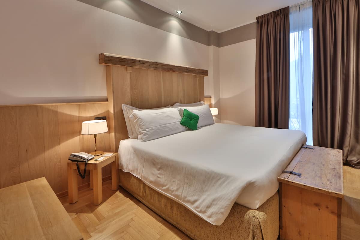 Habitación de hotel con una cama grande, cabecera de madera, lámparas de noche, una mesita de noche de madera y una ventana con cortinas.