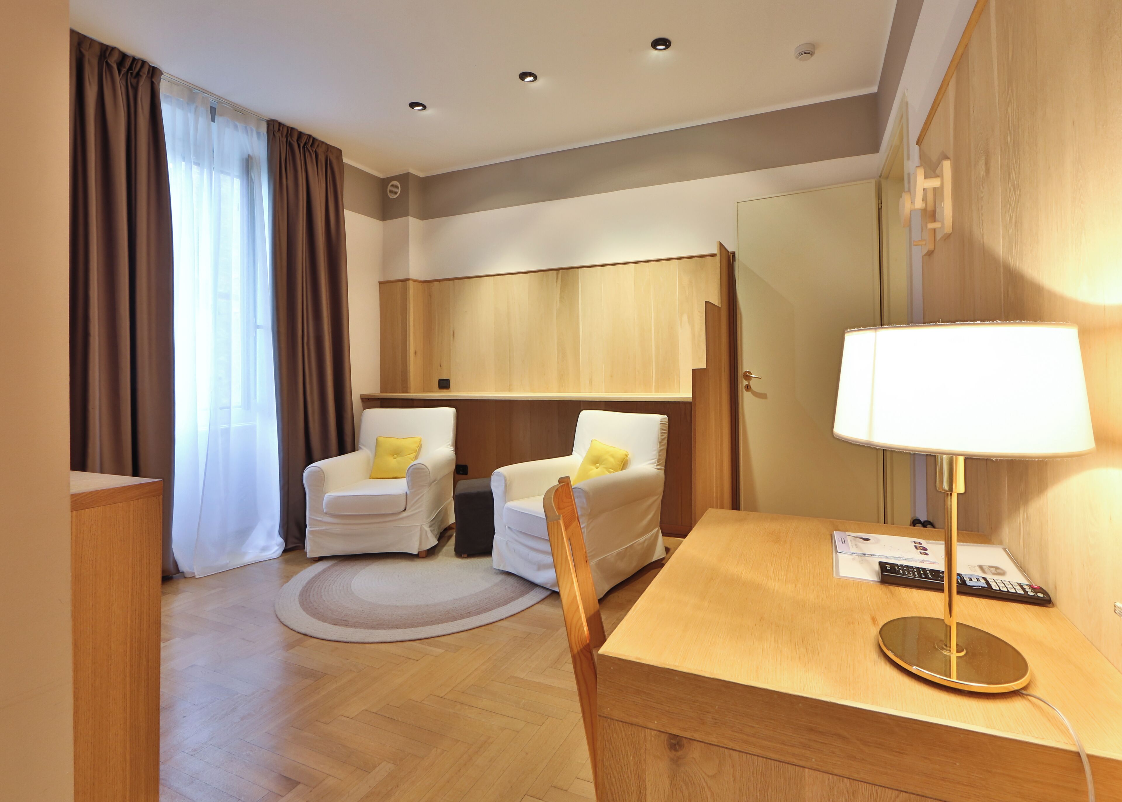 Habitación de hotel con dos sillones blancos, un escritorio de madera, una lámpara, un tapete redondo y un armario.