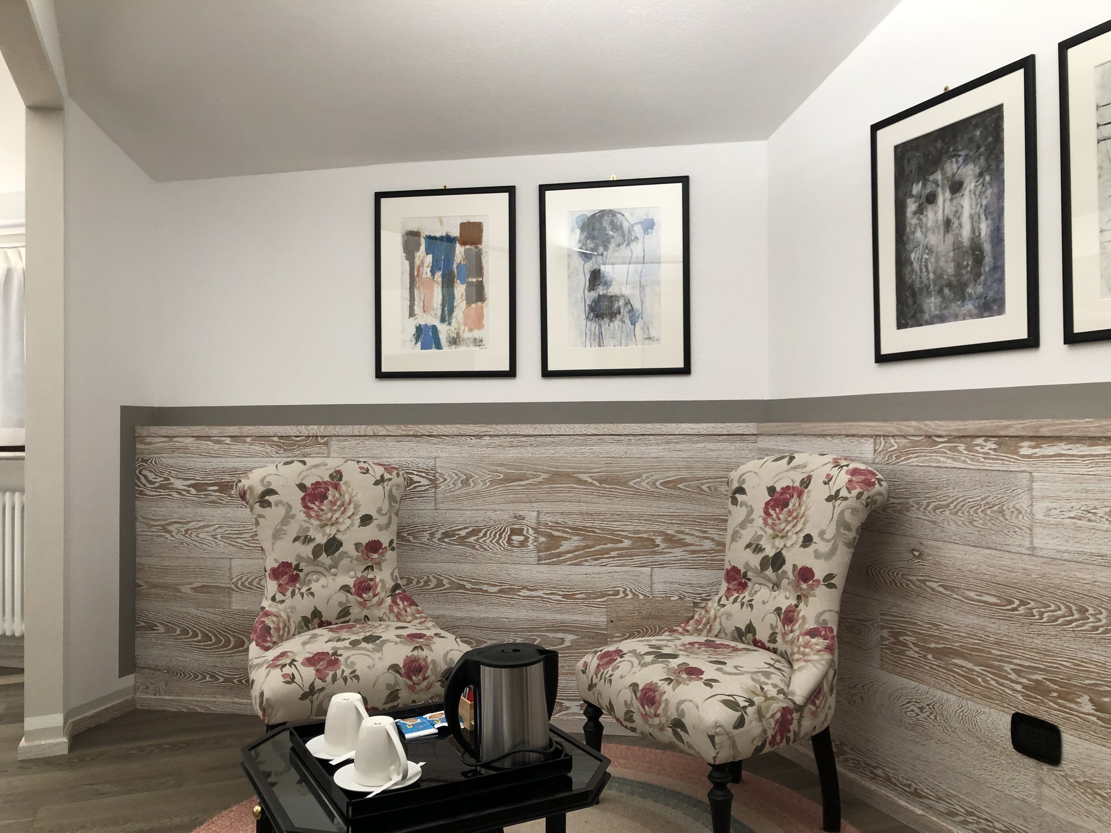 Una habitación con dos sillones tapizados con flores, una mesa de centro negra con una tetera y tazas, y tres cuadros abstractos enmarcados en la pared.
