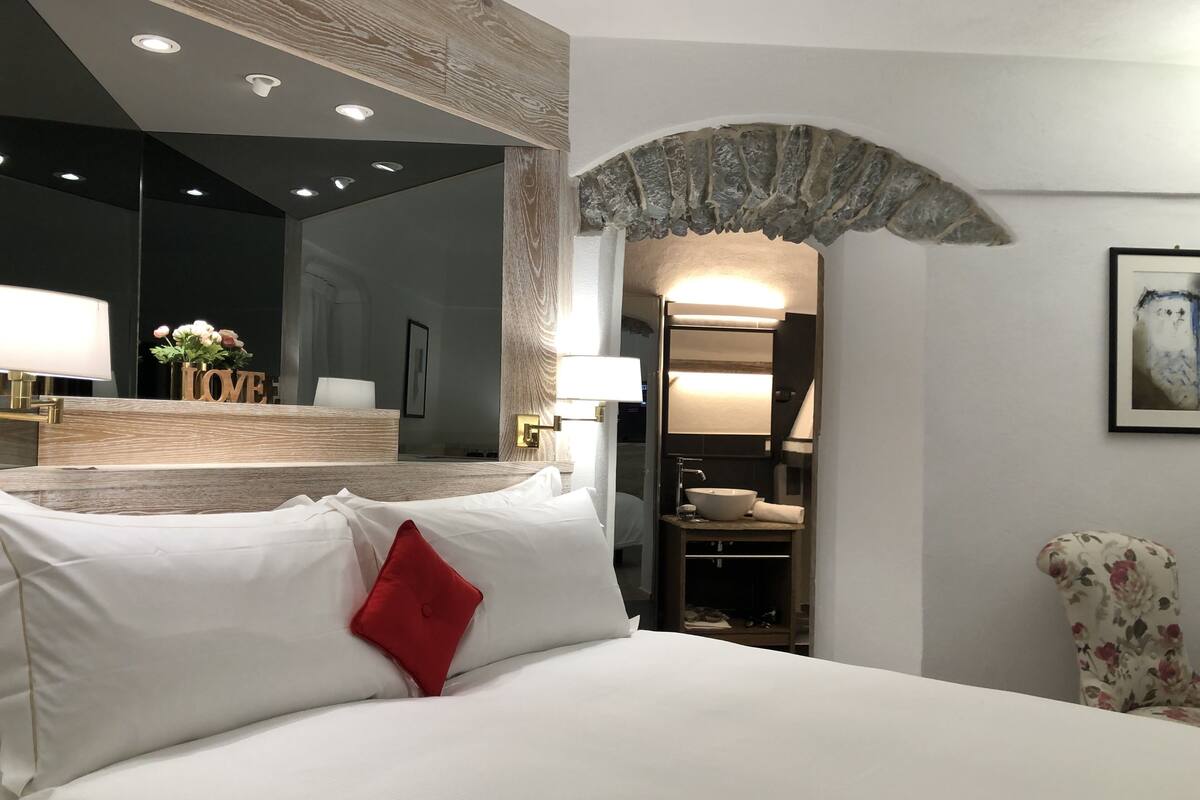 Habitación de hotel con una cama grande, cabecera de madera, un espejo, tocador con lavabo y una obra de arte enmarcada en la pared.