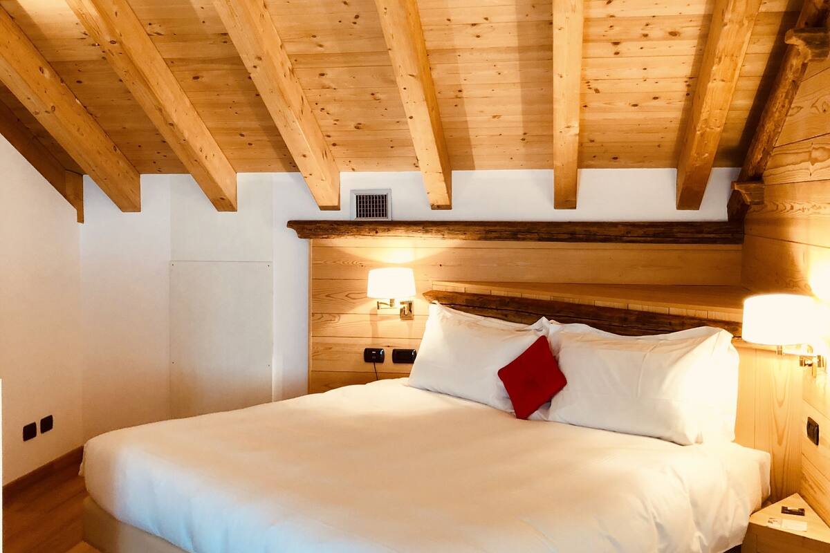 Un dormitorio con techo de madera, cama con ropa blanca, almohada roja y lámparas de noche.