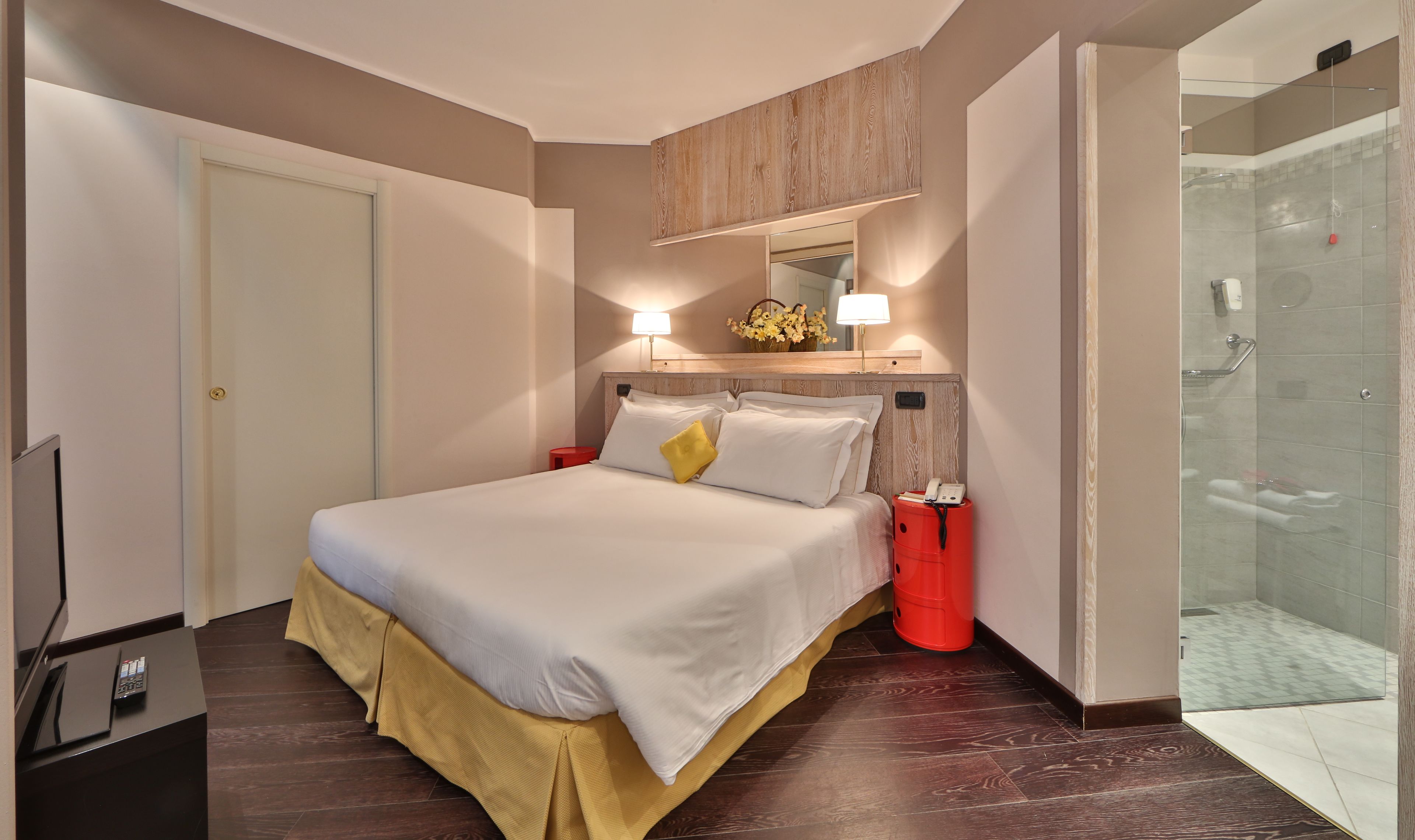 Habitación de hotel con cama, mesitas de noche, espejo, zona de ducha y televisión.