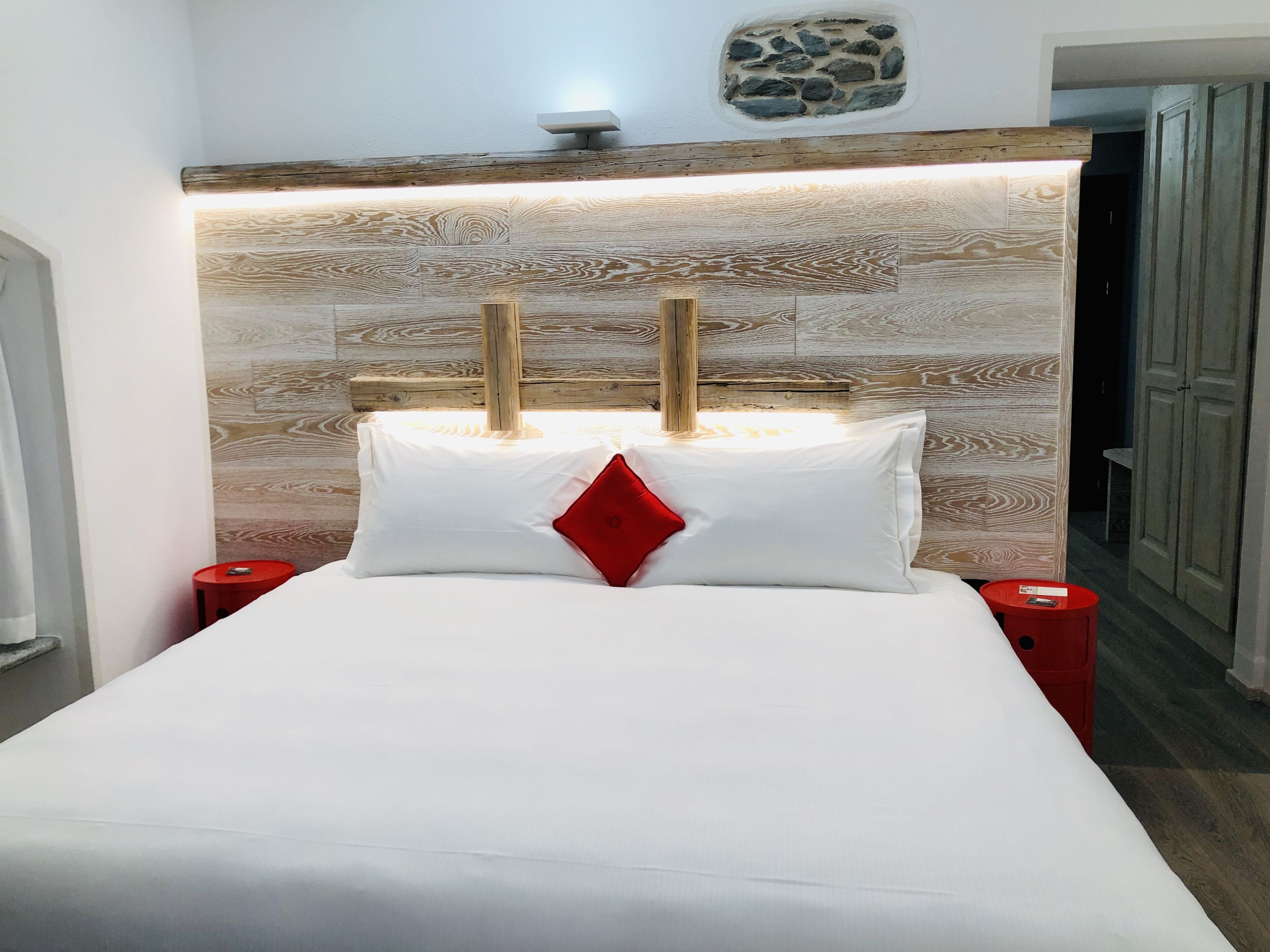 Una cama con ropa de cama blanca y almohada roja, cabecera de madera con detalle de piedra y dos mesitas de noche rojas.