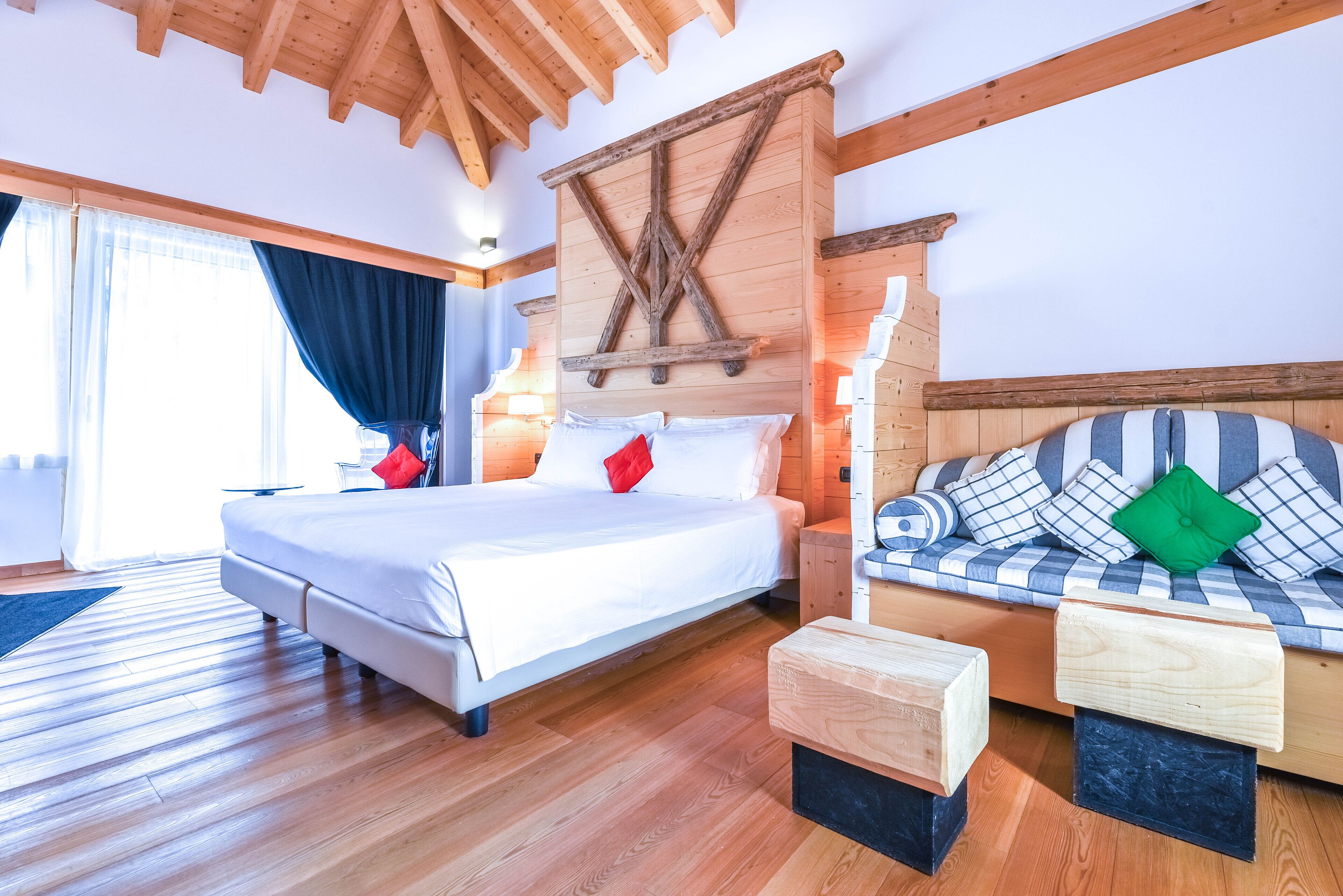 A bedroom with a wooden ceiling, a bed with white bedding, a sofa with patterned cushions, and a wooden nightstand.