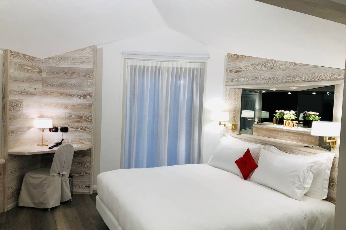Habitación de hotel con cama, escritorio, silla, ventana con cortinas y espejo con marco decorativo.
