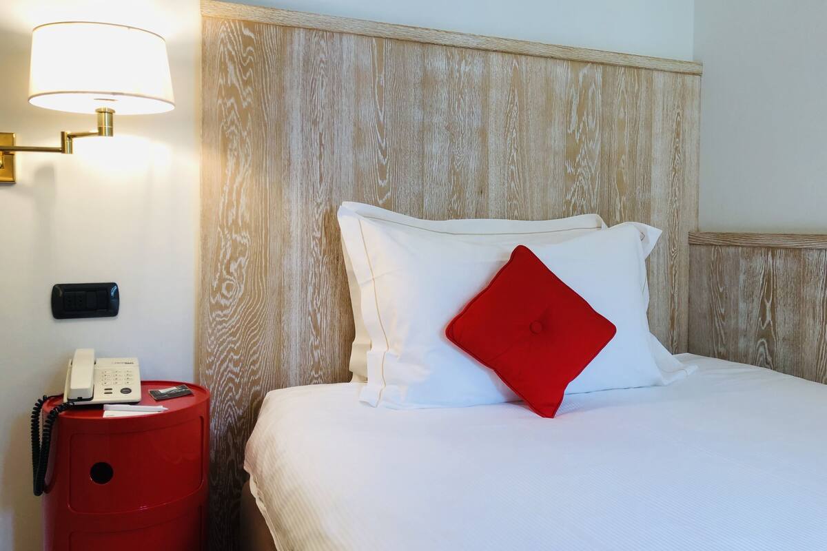 Habitación de hotel con cabecera de madera, cama blanca con almohada roja, mesita de noche con lámpara y cesto de basura rojo.