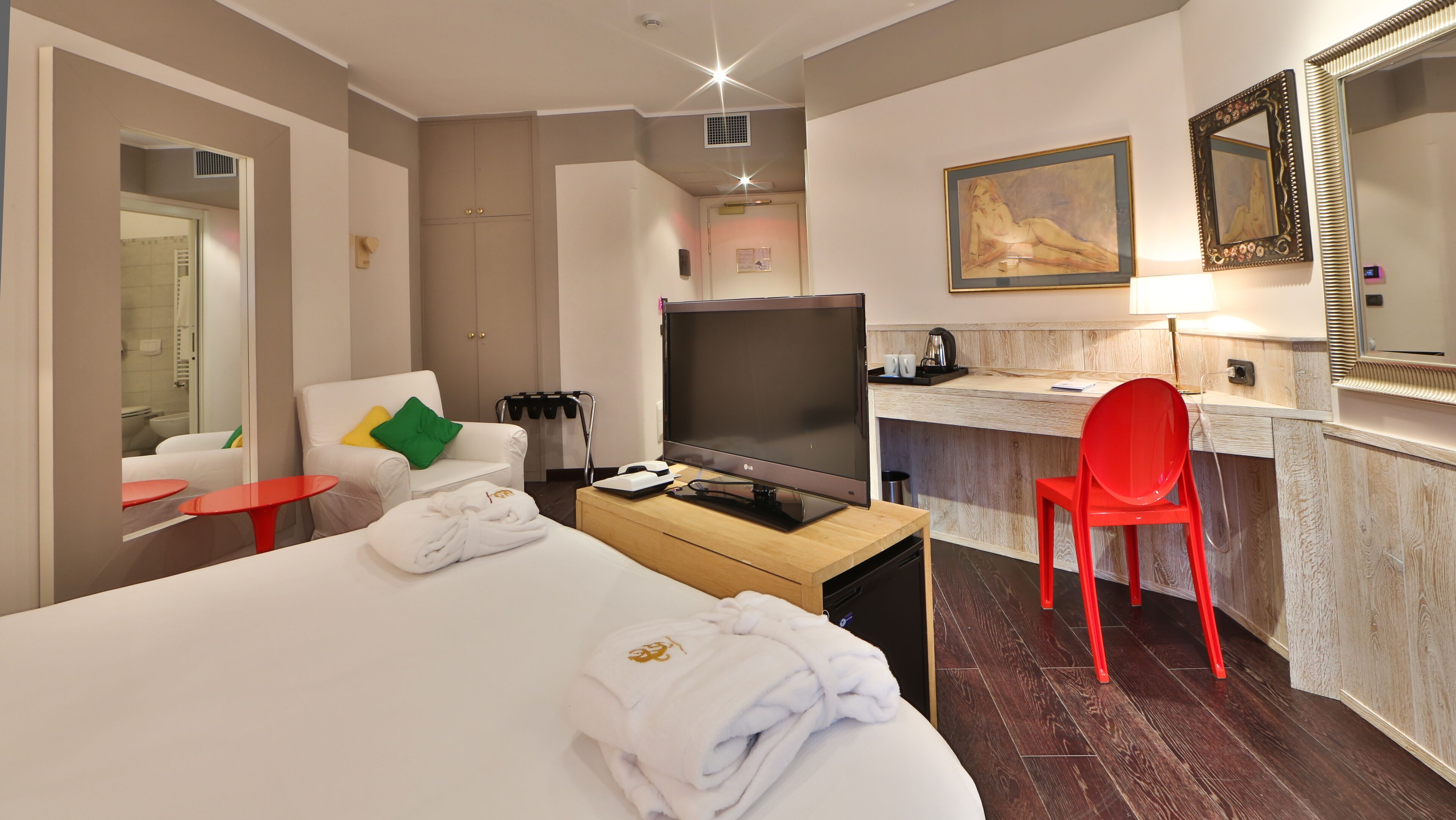 Une chambre d’hôtel avec un lit, un bureau avec un ordinateur, une télévision, un fauteuil rouge et un tableau au mur.