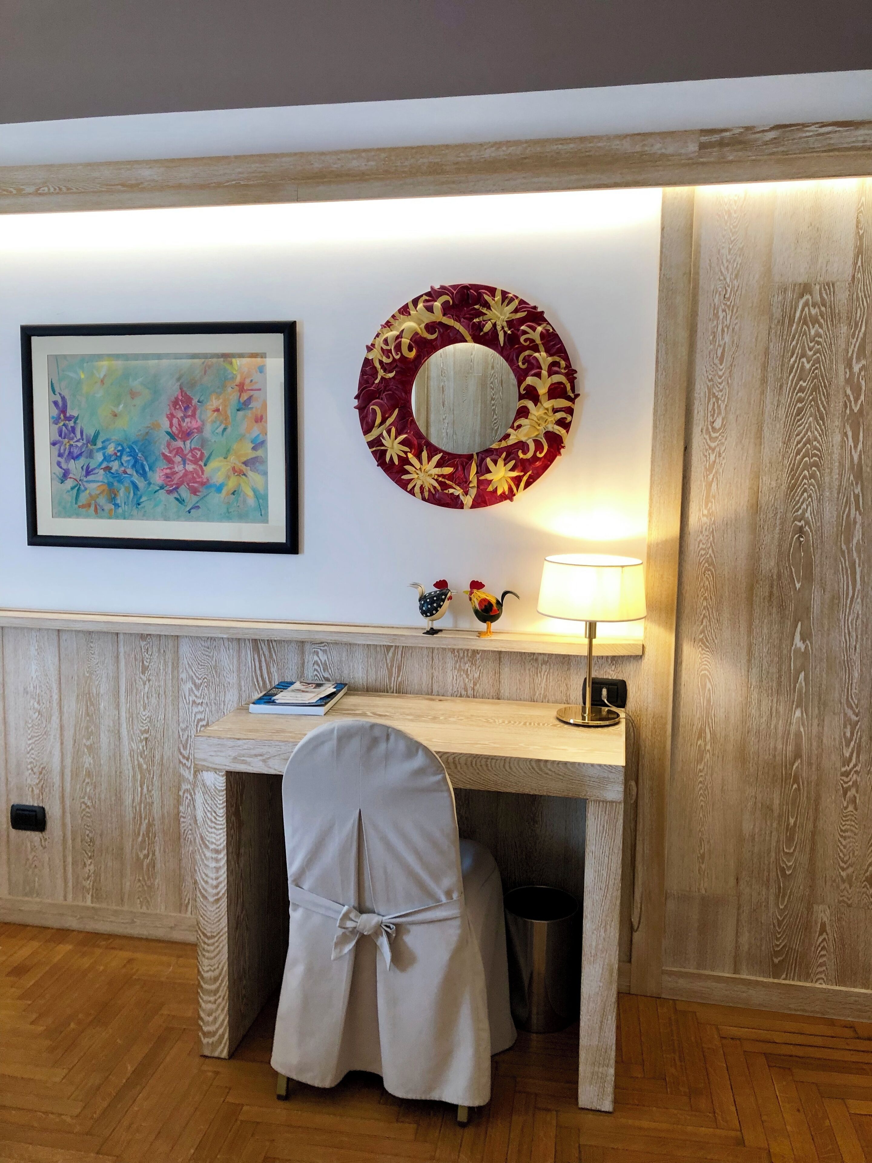 A wooden desk with a chair, a framed painting, a decorative mirror, and a lamp.