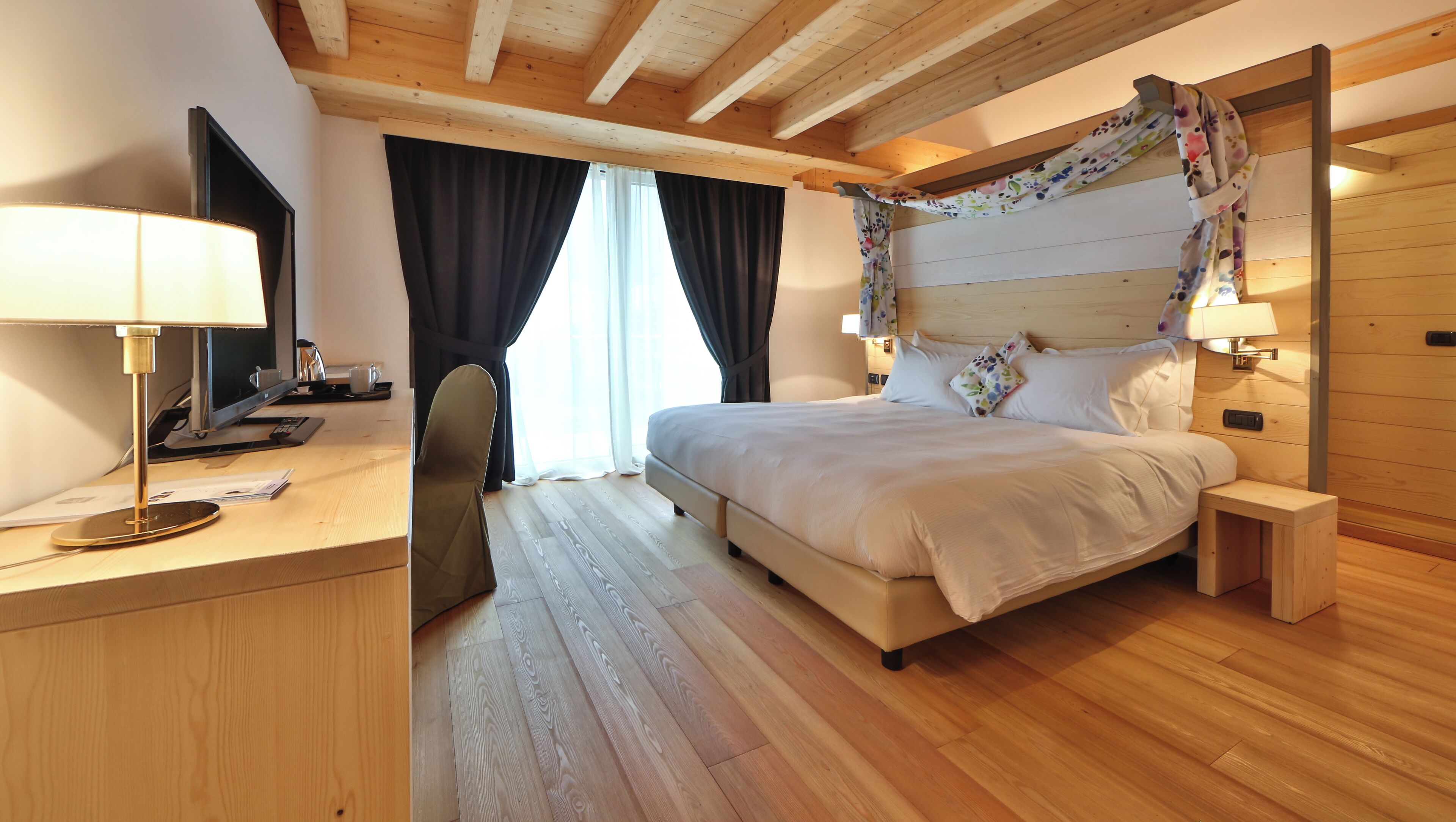 A bedroom with a wooden ceiling, a bed with white bedding, a desk with a lamp, and a chair.