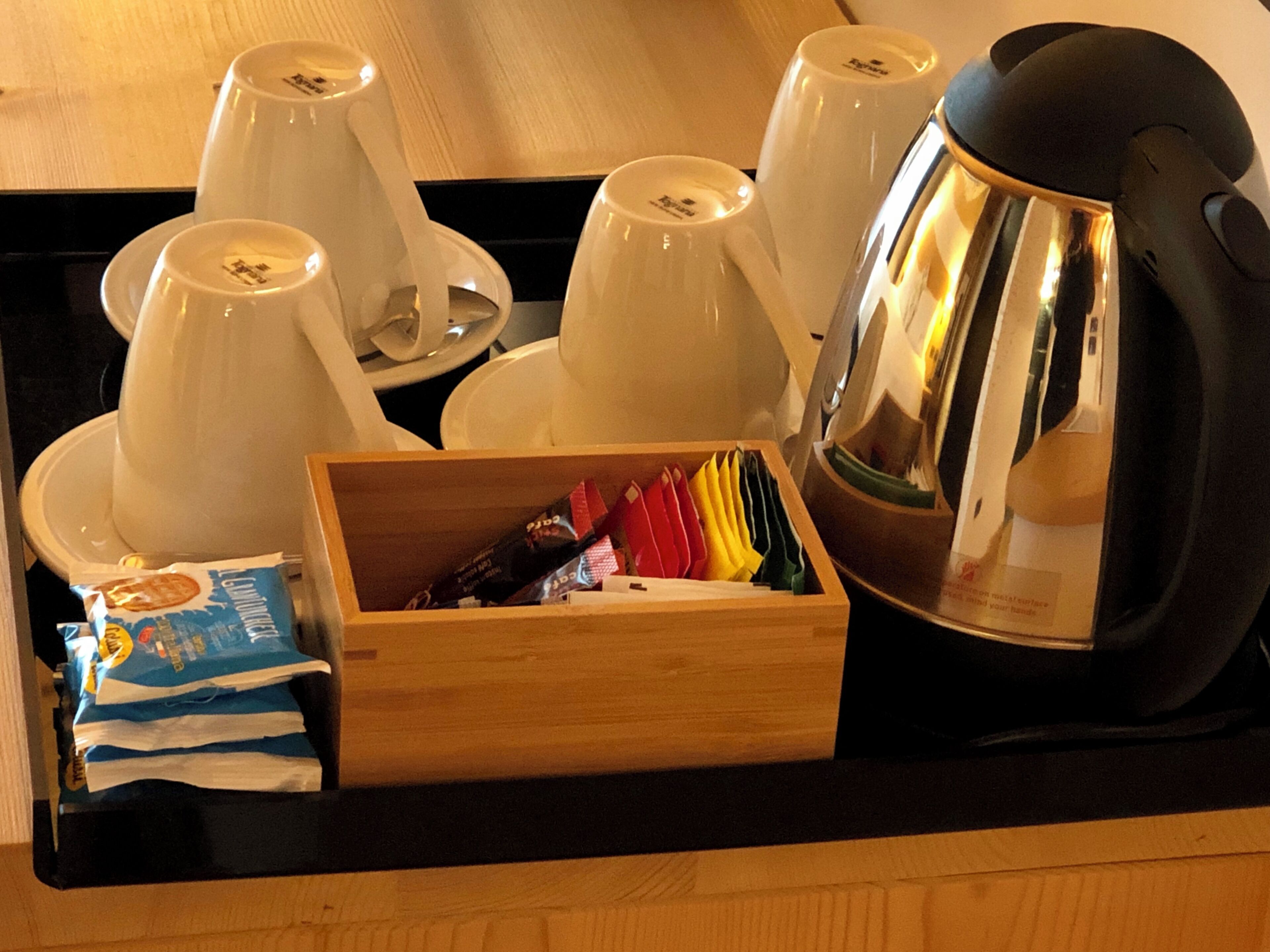 Una bandeja con tazas blancas, una cafetera negra y una caja de madera que contiene servilletas de colores y un paquete de pañuelos.