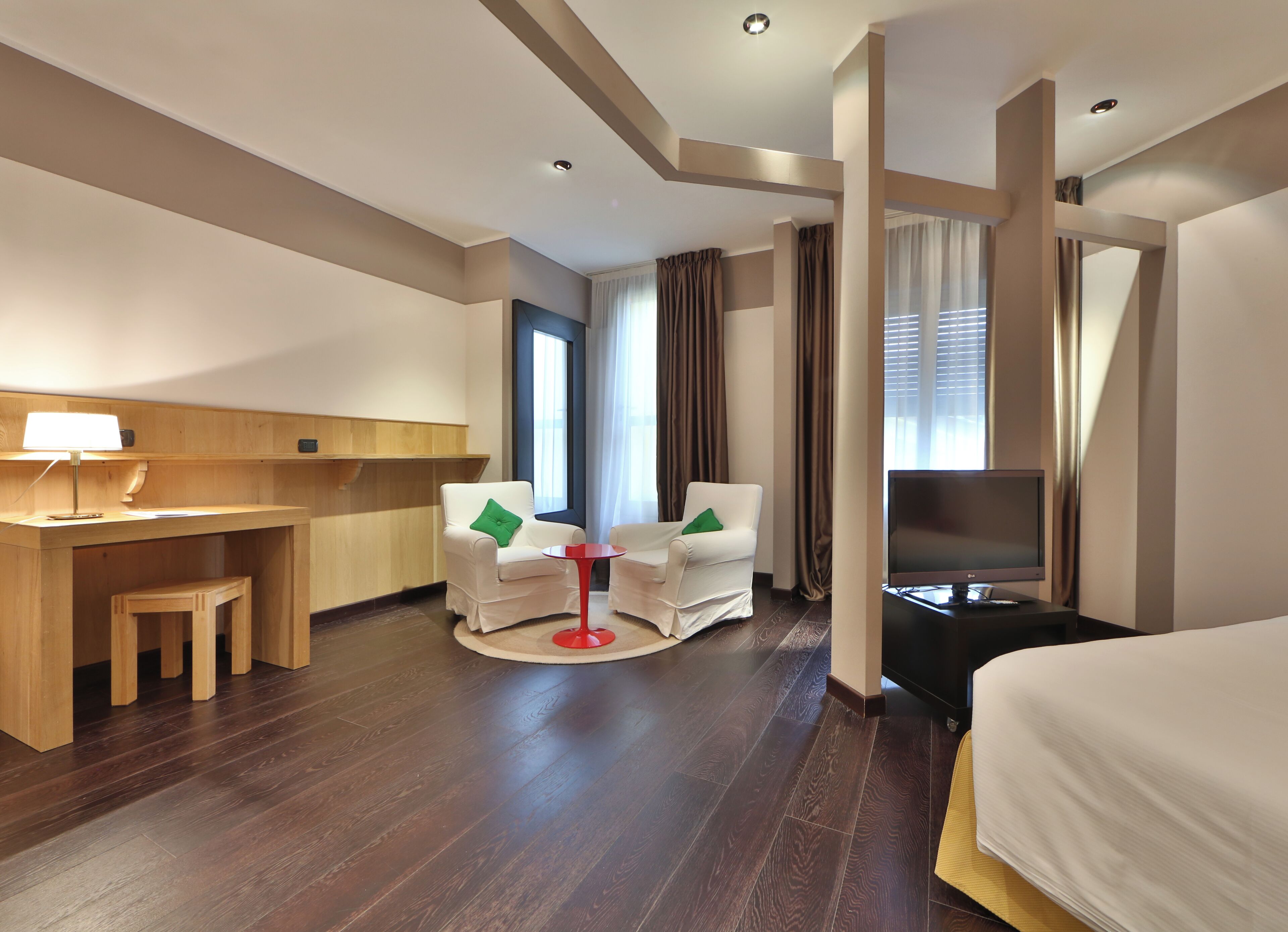 Habitación de hotel con un escritorio de madera, dos sillones blancos con cojines verdes, una mesita roja pequeña, un televisor de pantalla plana y una cama con edredón blanco.