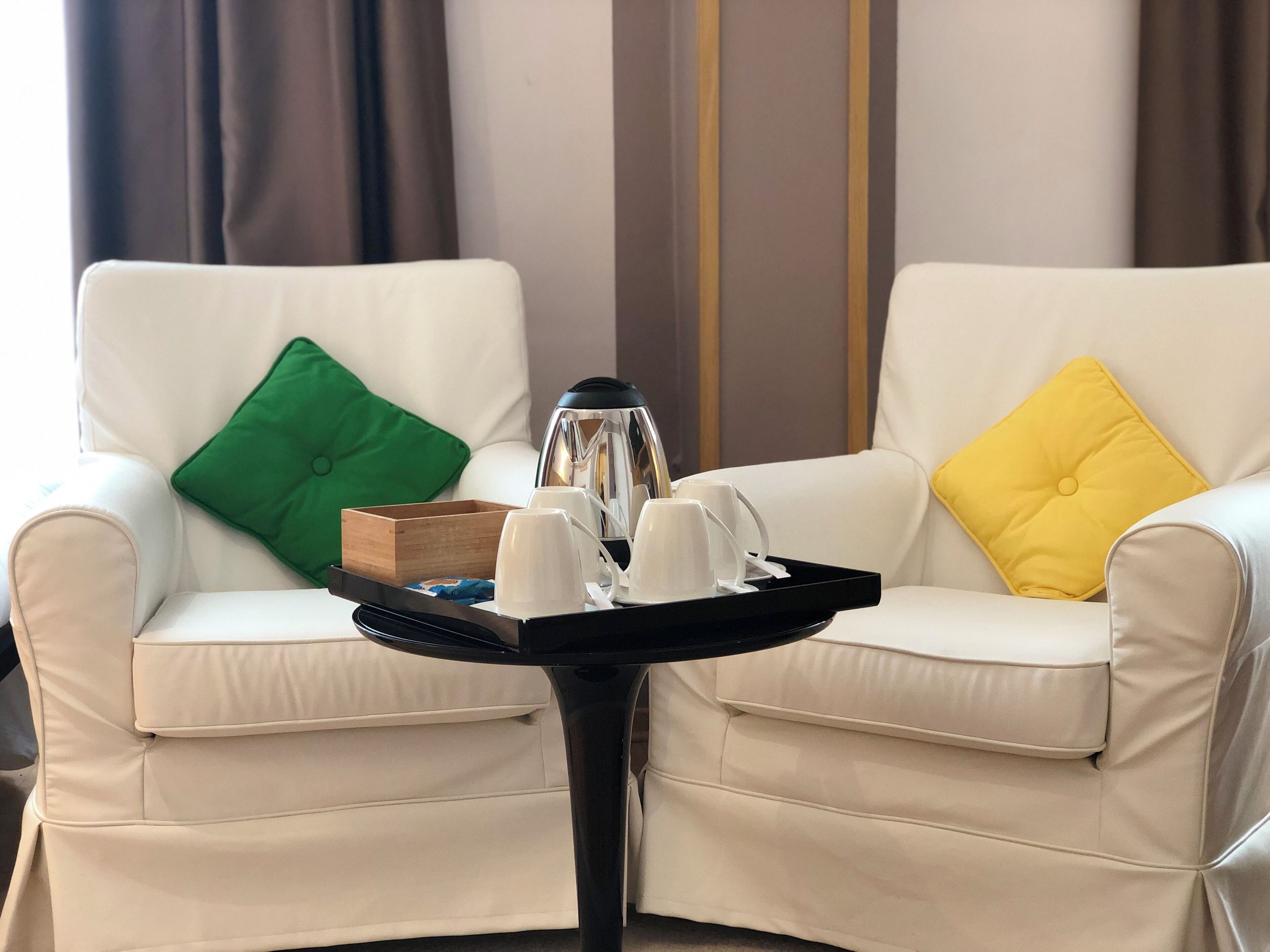 A hotel room with two white leather armchairs, each with a colorful cushion, a small wooden tray with a teapot, cups, and a book, and a black side table.