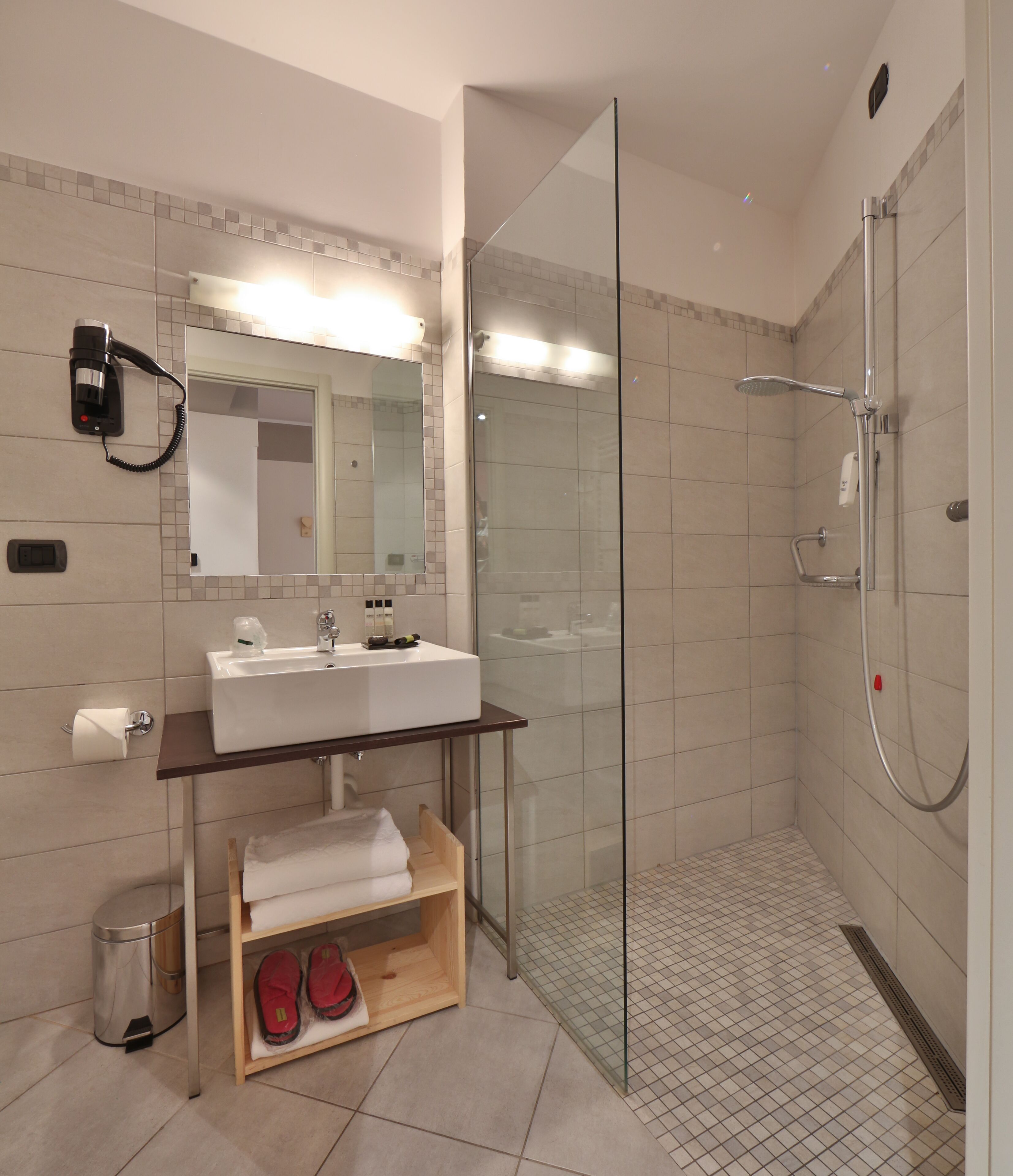 A modern bathroom with a glass-enclosed shower, a sink with a mirror above, and a shelf with towels and slippers.