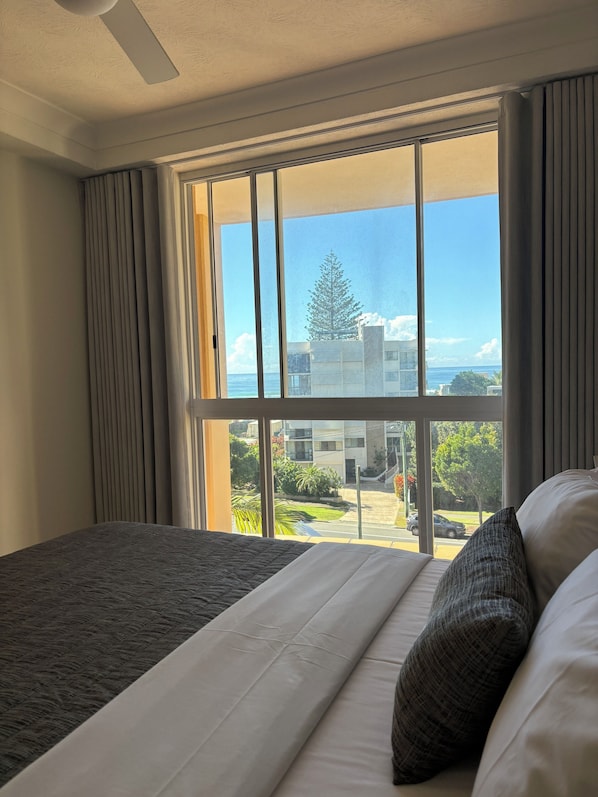 A hotel room with a large window, a bed with white and grey bedding, and a view of a building and greenery outside.