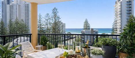 A balcony with a table set for a meal, overlooking a coastal view with buildings and trees.