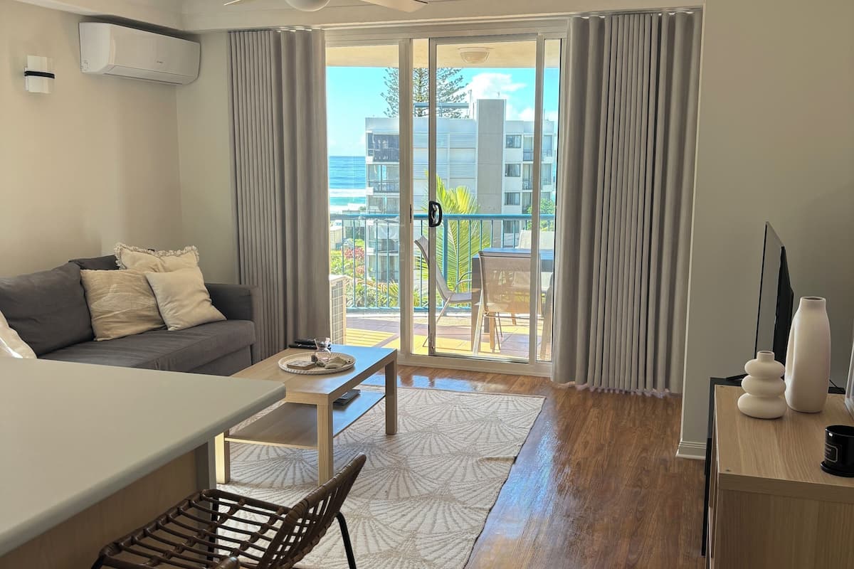 A modern living room with a sofa, coffee table, and a dining area. There is a balcony with a view of buildings and the ocean.
