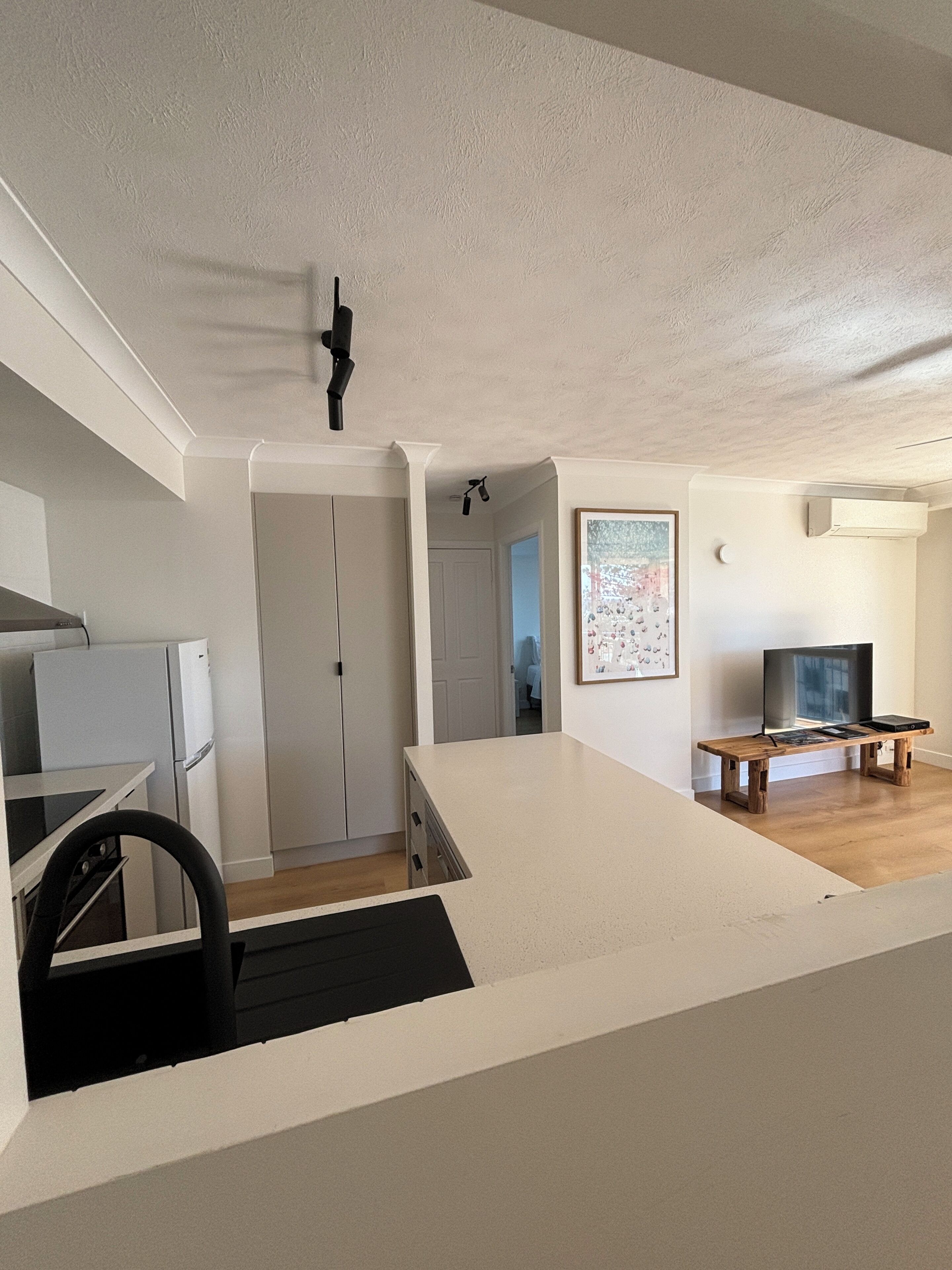 Une cuisine moderne avec des armoires blanches, un four encastré et un comptoir noir. On y trouve un banc en bois, un téléviseur et un tableau encadré accroché au mur.