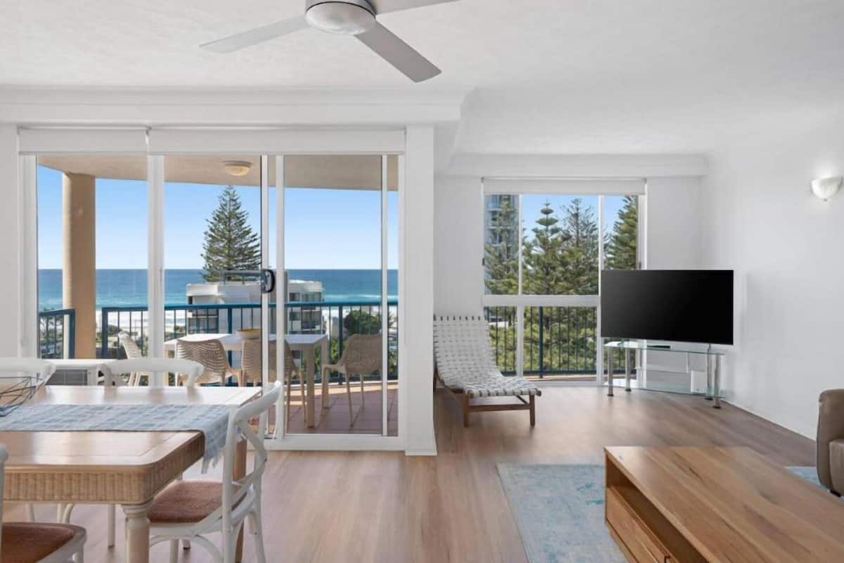 A spacious living room with a balcony overlooking the ocean, a flat-screen TV, and a dining area with wicker furniture.